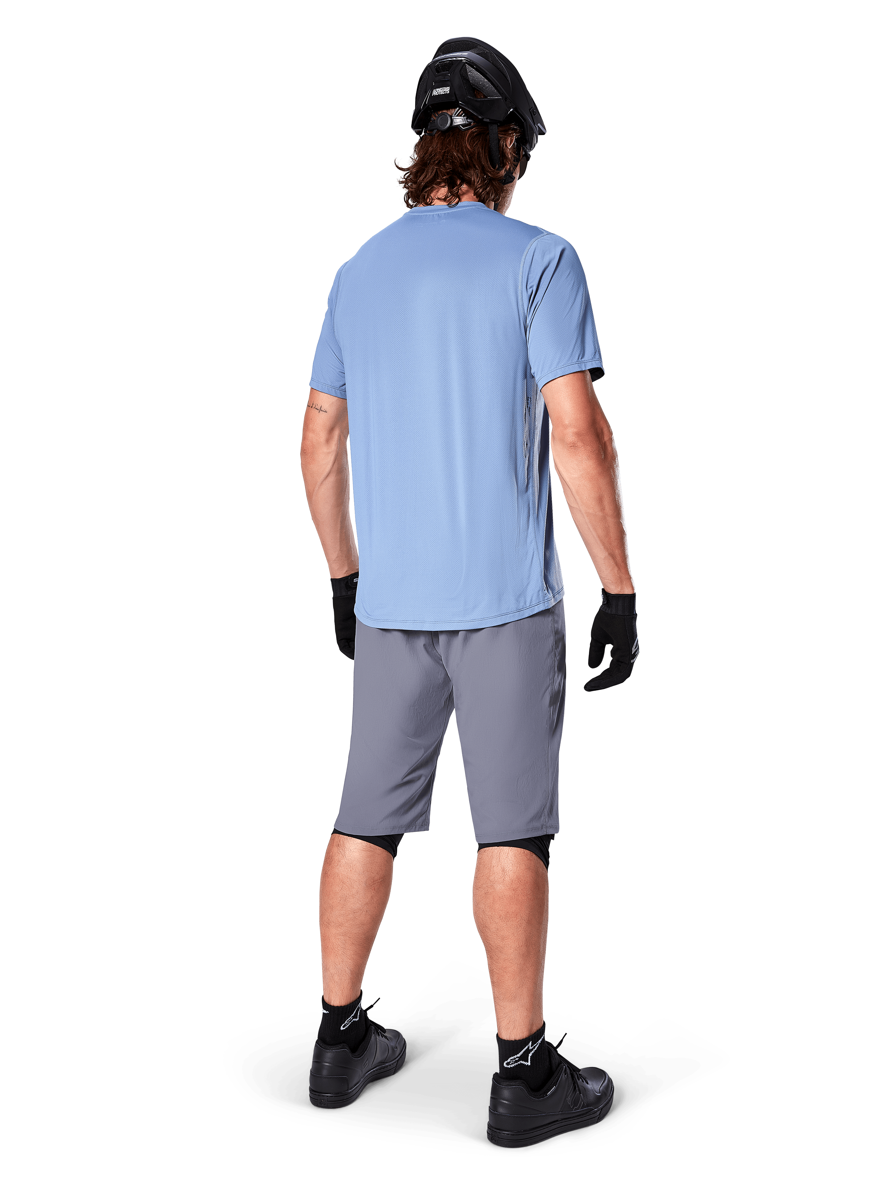 Alpinestars A-Dura Shorts, pantaloncini MTB grigio scuro, vista posteriore, indossati con maglia azzurra a maniche corte e casco da mountain bike nero, guanti e calze neri con scarpe nere, abbigliamento funzionale e resistente per il ciclismo off-road