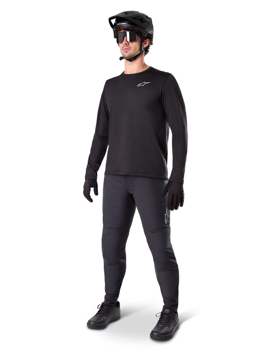 Modello con maglia termica Alpinestars A-Dura, maglia da MTB a maniche lunghe nera, casco da mountain bike nero, occhiali da sole e pantaloni grigio scuro, caratterizzata da un piccolo logo bianco sul petto, struttura in pile isolante per uscite con il freddo