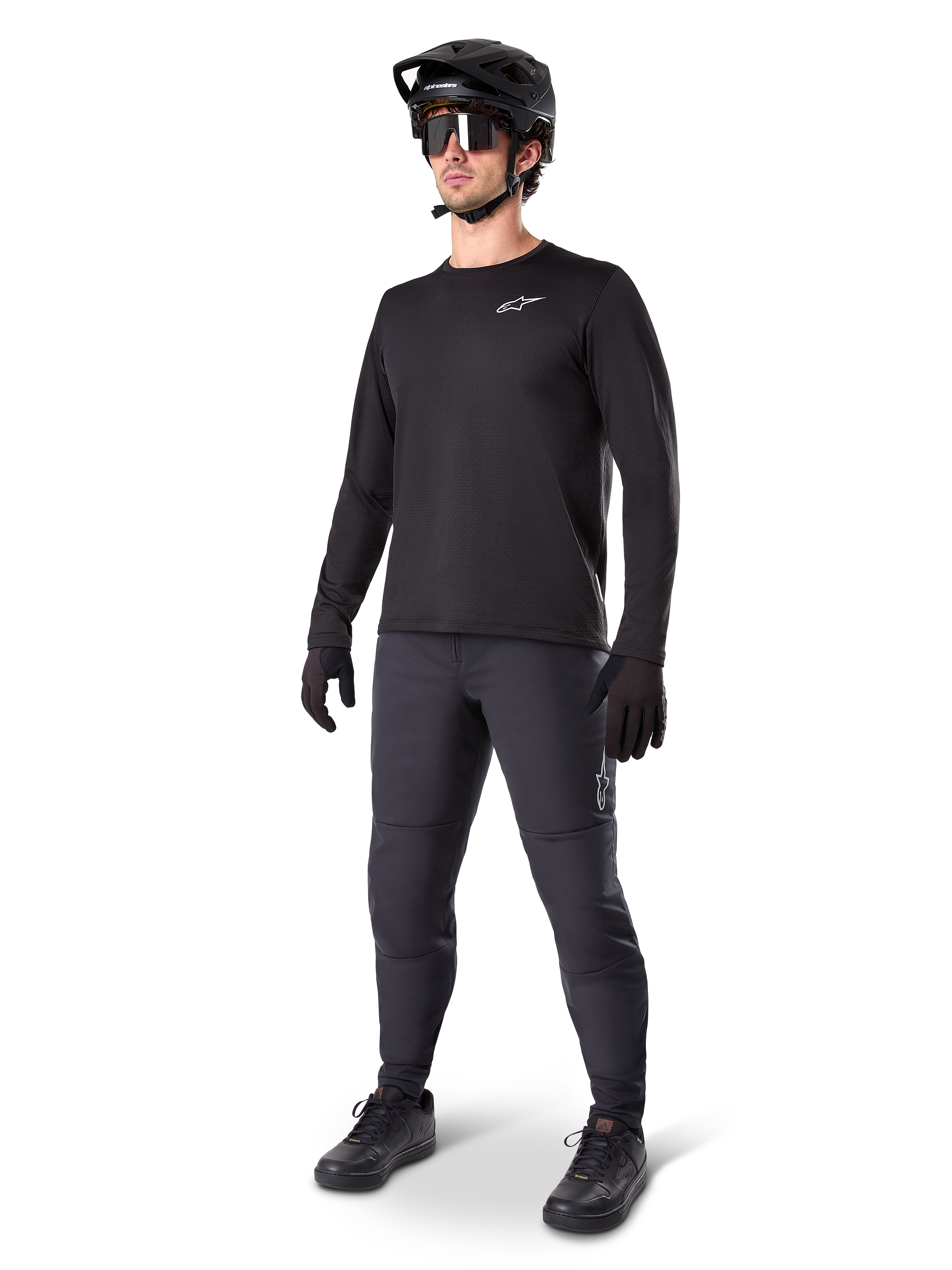 Modello con maglia termica Alpinestars A-Dura, maglia da MTB a maniche lunghe nera, casco da mountain bike nero, occhiali da sole e pantaloni grigio scuro, caratterizzata da un piccolo logo bianco sul petto, struttura in pile isolante per uscite con il freddo