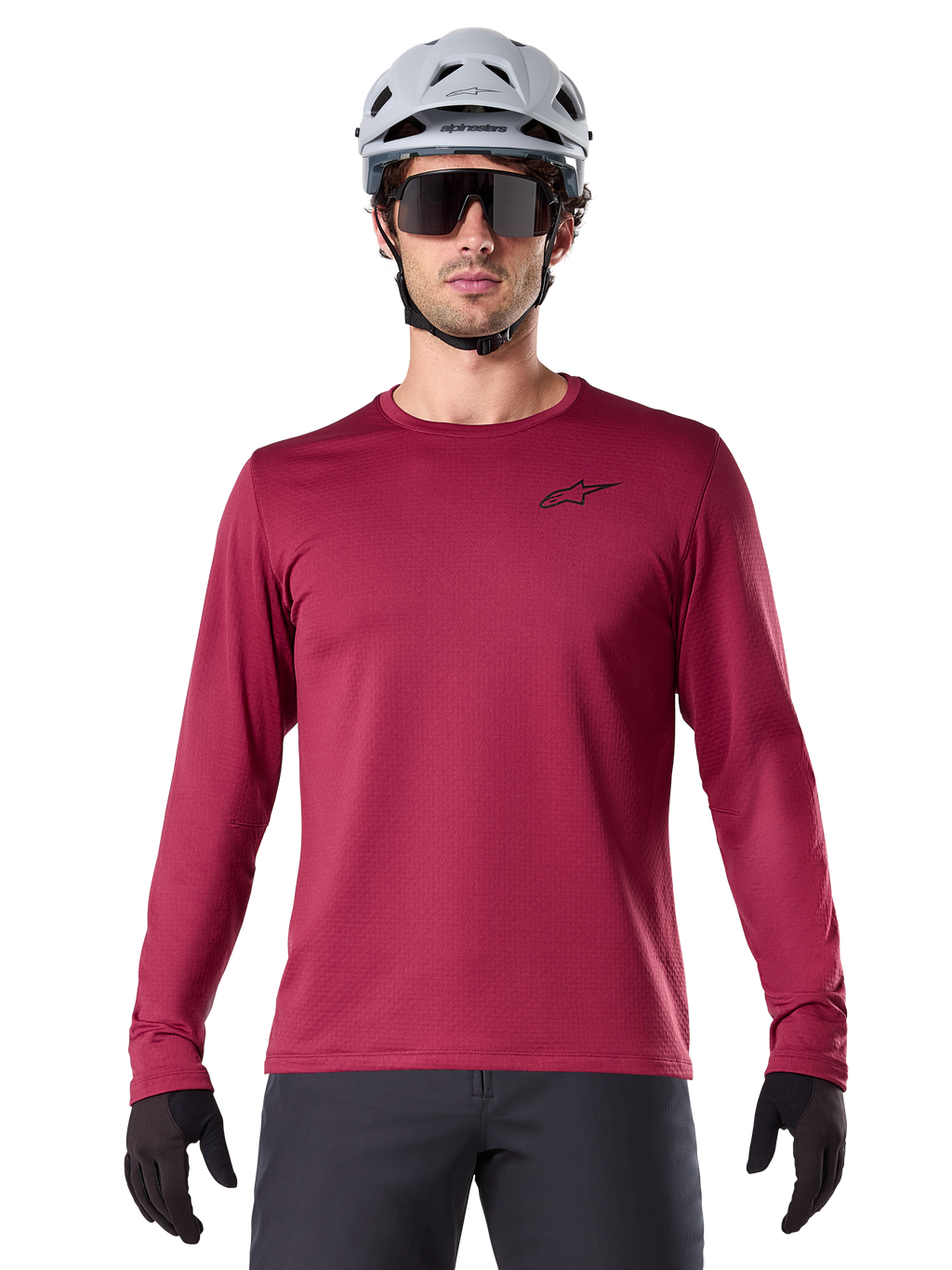 Ciclista uomo che indossa la maglia termica Alpinestars A-Dura, maglia MTB a maniche lunghe bordeaux, abbinata a un casco grigio e occhiali da sole neri, piccolo logo a stella nero sul petto, tessuto in pile isolante per mountain bike in climi freddi