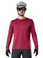 Ciclista uomo che indossa la maglia termica Alpinestars A-Dura, maglia MTB a maniche lunghe bordeaux, abbinata a un casco grigio e occhiali da sole neri, piccolo logo a stella nero sul petto, tessuto in pile isolante per mountain bike in climi freddi