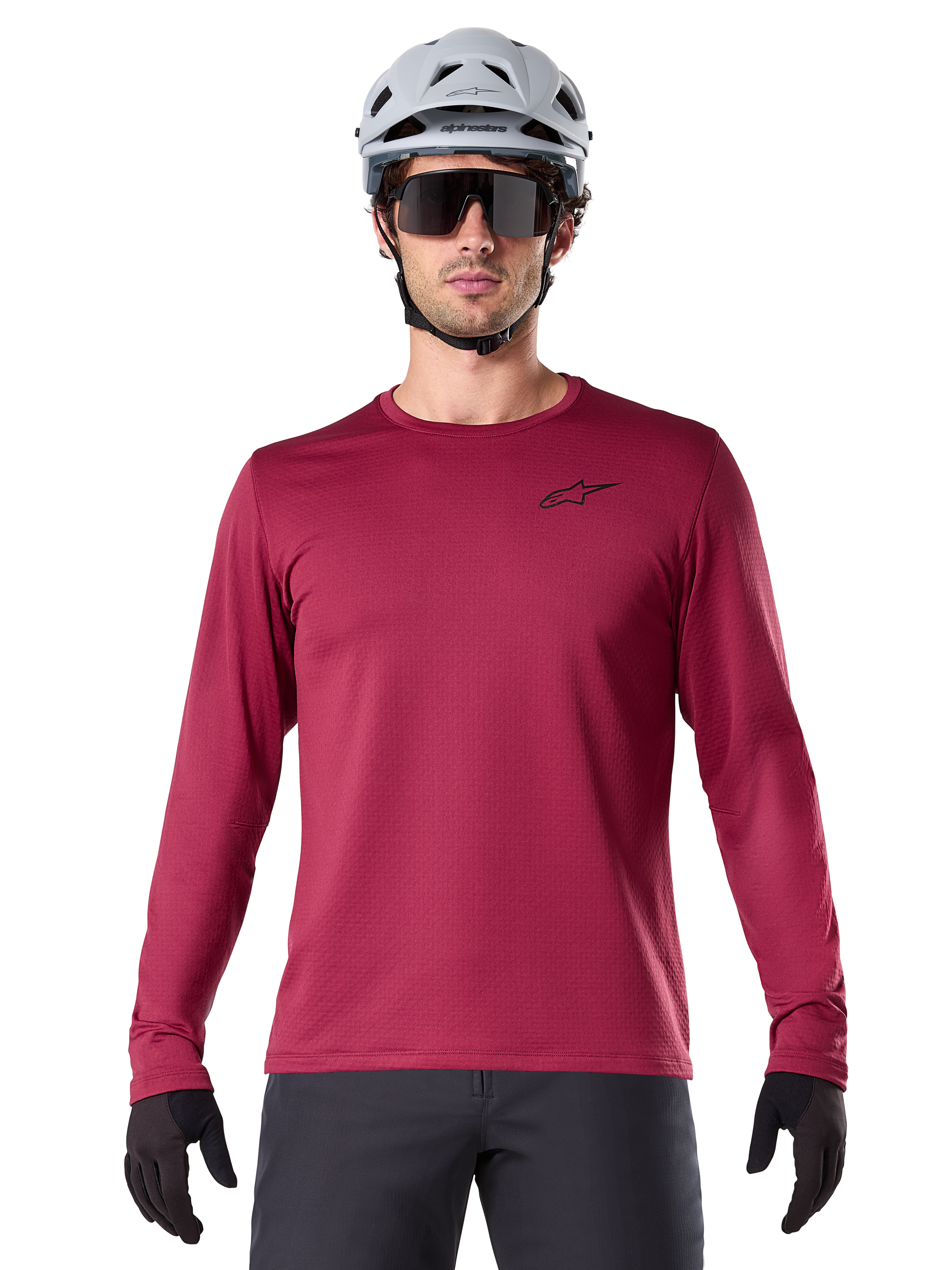 Ciclista uomo che indossa la maglia termica Alpinestars A-Dura, maglia MTB a maniche lunghe bordeaux, abbinata a un casco grigio e occhiali da sole neri, piccolo logo a stella nero sul petto, tessuto in pile isolante per mountain bike in climi freddi