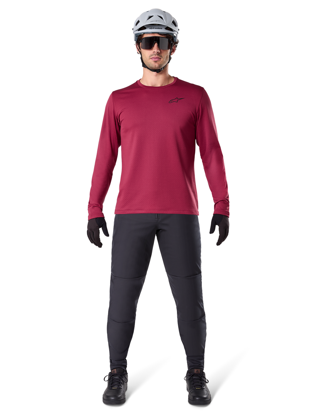 Modello uomo che indossa la maglia termica Alpinestars A-Dura colore Burgundy, maglia da ciclismo isolante a maniche lunghe, abbinata a pantaloni da mountain bike grigio scuro, casco bianco, occhiali da sole neri e guanti neri, equipaggiamento professionale da MTB per il freddo