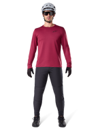 Modello uomo che indossa la maglia termica Alpinestars A-Dura colore Burgundy, maglia da ciclismo isolante a maniche lunghe, abbinata a pantaloni da mountain bike grigio scuro, casco bianco, occhiali da sole neri e guanti neri, equipaggiamento professionale da MTB per il freddo