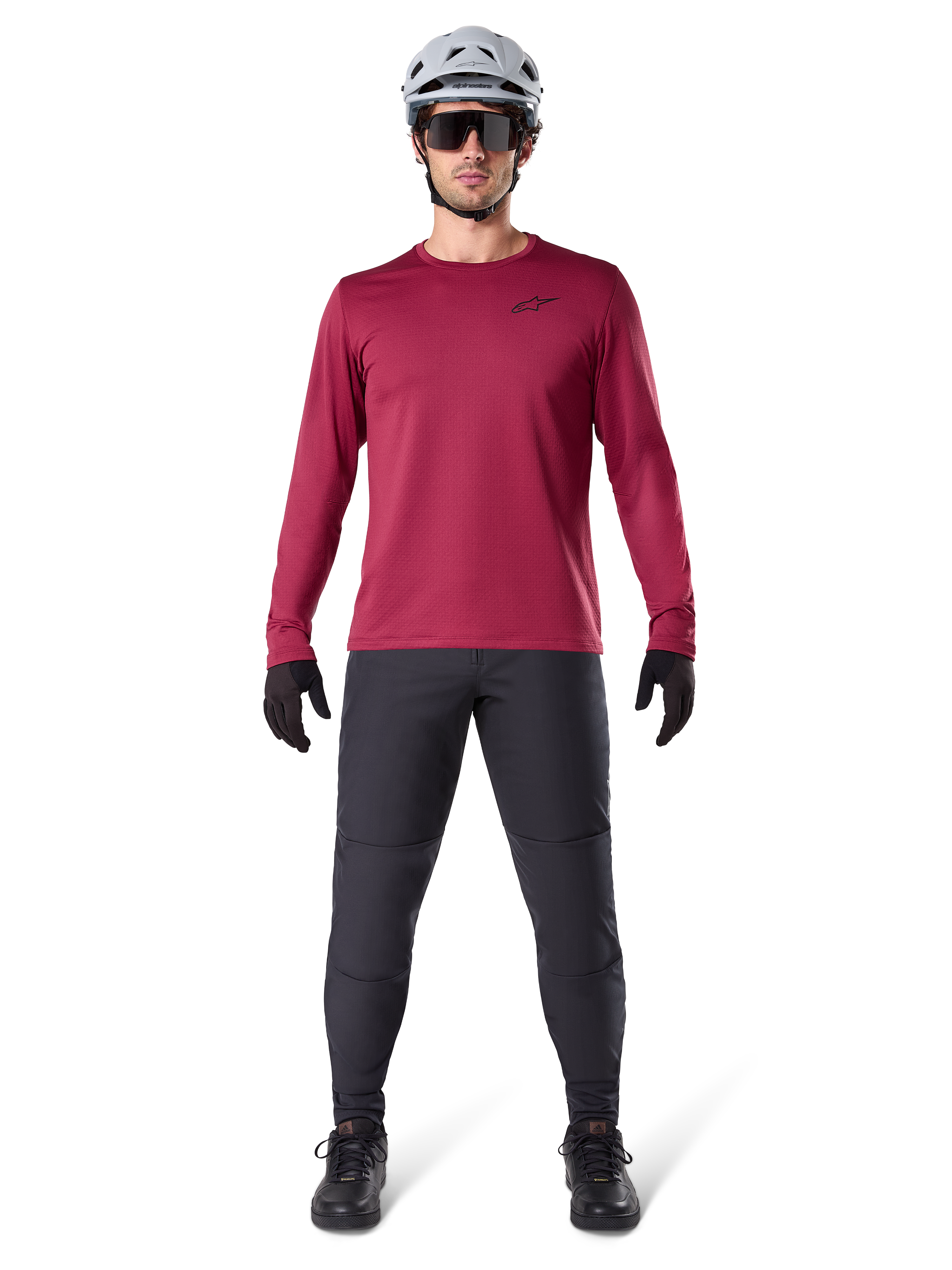 Modello uomo che indossa la maglia termica Alpinestars A-Dura colore Burgundy, maglia da ciclismo isolante a maniche lunghe, abbinata a pantaloni da mountain bike grigio scuro, casco bianco, occhiali da sole neri e guanti neri, equipaggiamento professionale da MTB per il freddo