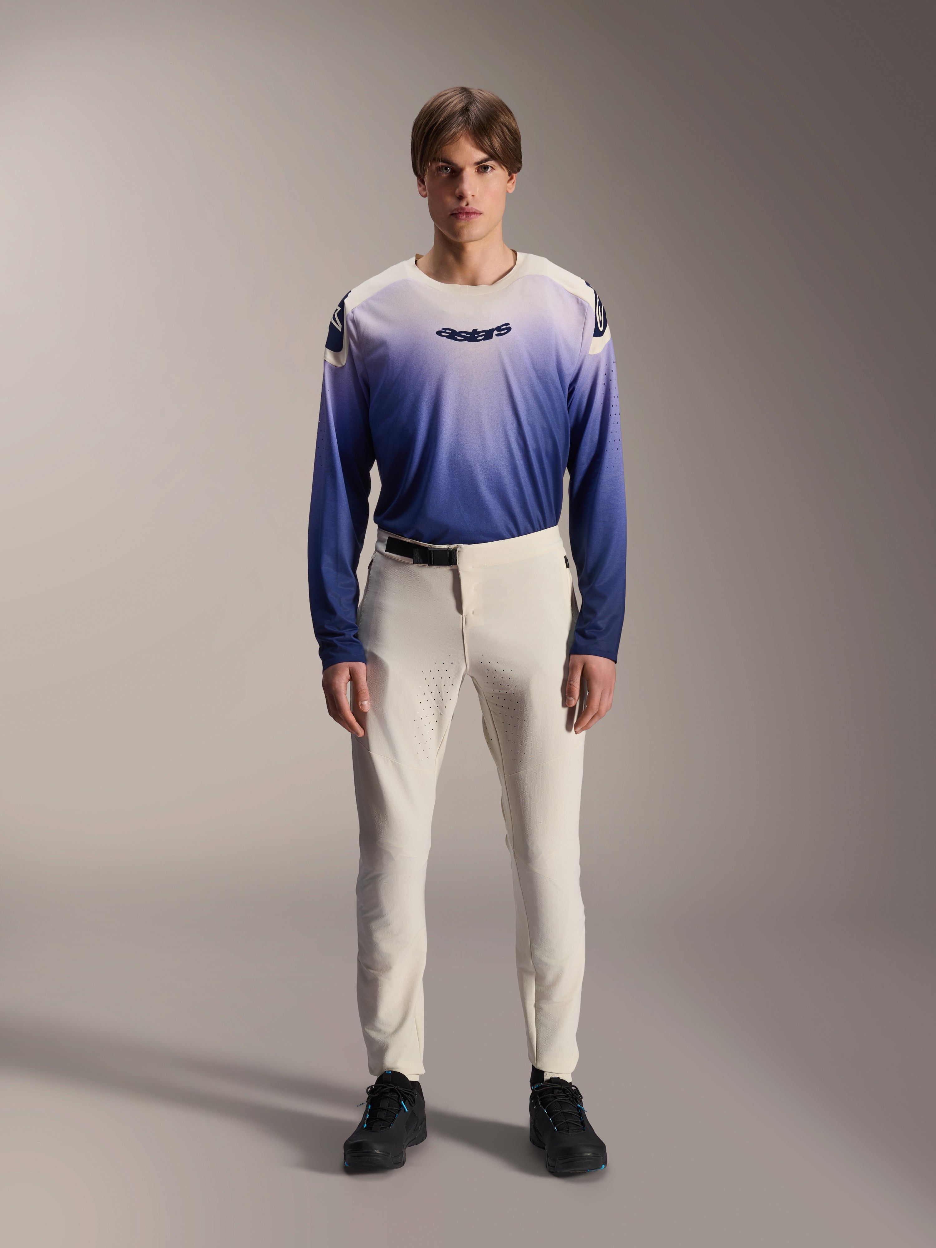 Alpinestars A-Aria Elite Pants, pantaloni da MTB performance in colore Ecru, pantaloni da ciclismo bianco panna con pannelli sulle cosce traforati al laser e fibbia magnetica, indossati con maglia a maniche lunghe blu sfumata e scarpe nere, vestibilità tecnica per trail riding