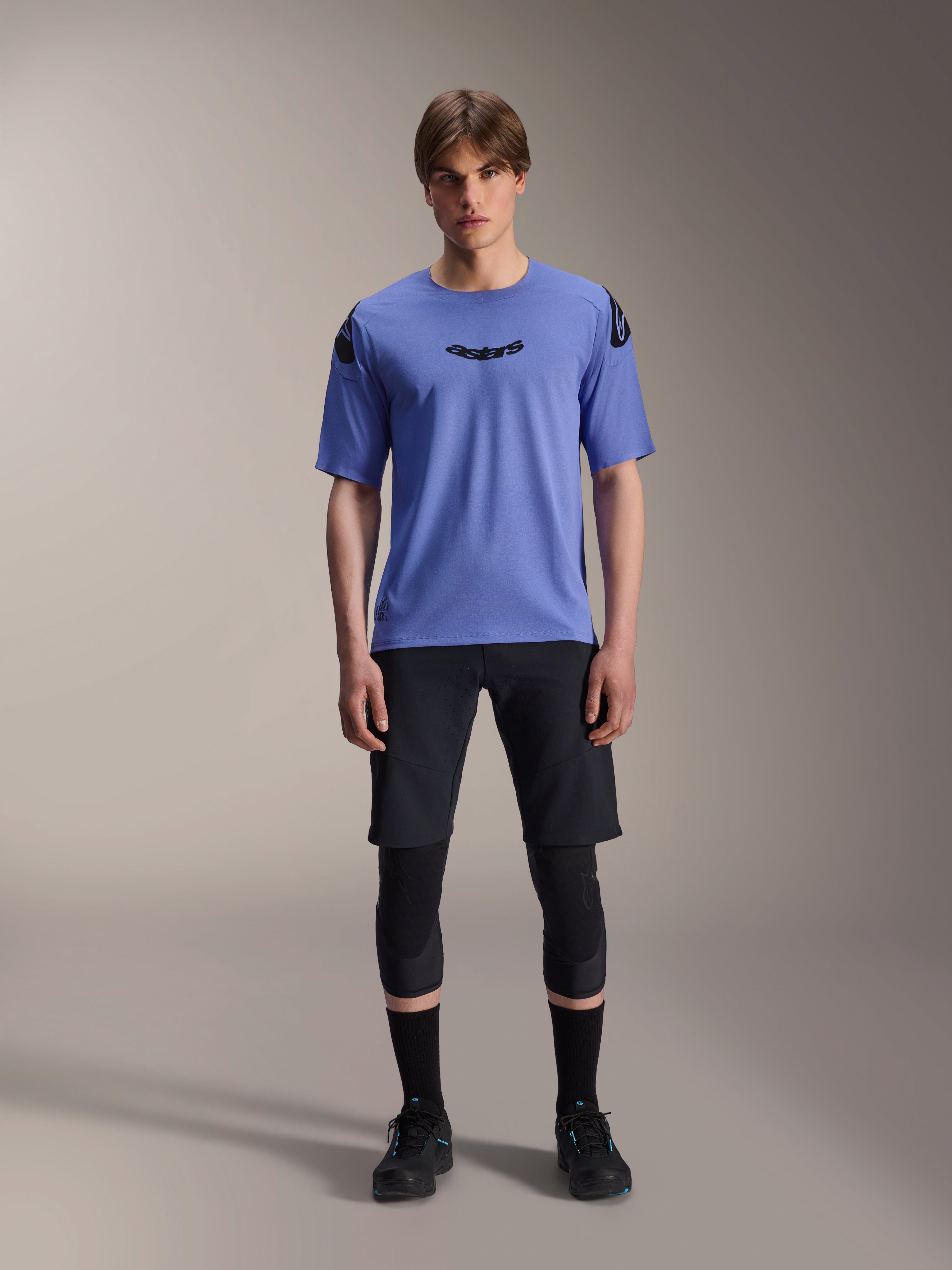 Alpinestars A-Aria Elite Shorts, pantaloncini da mountain bike MTB, Nero Titanium, indossati con maglia blu a maniche corte, protezioni per le ginocchia e scarpe da ciclismo nere, scatto in studio a figura intera, abbigliamento tecnico per mountain bike