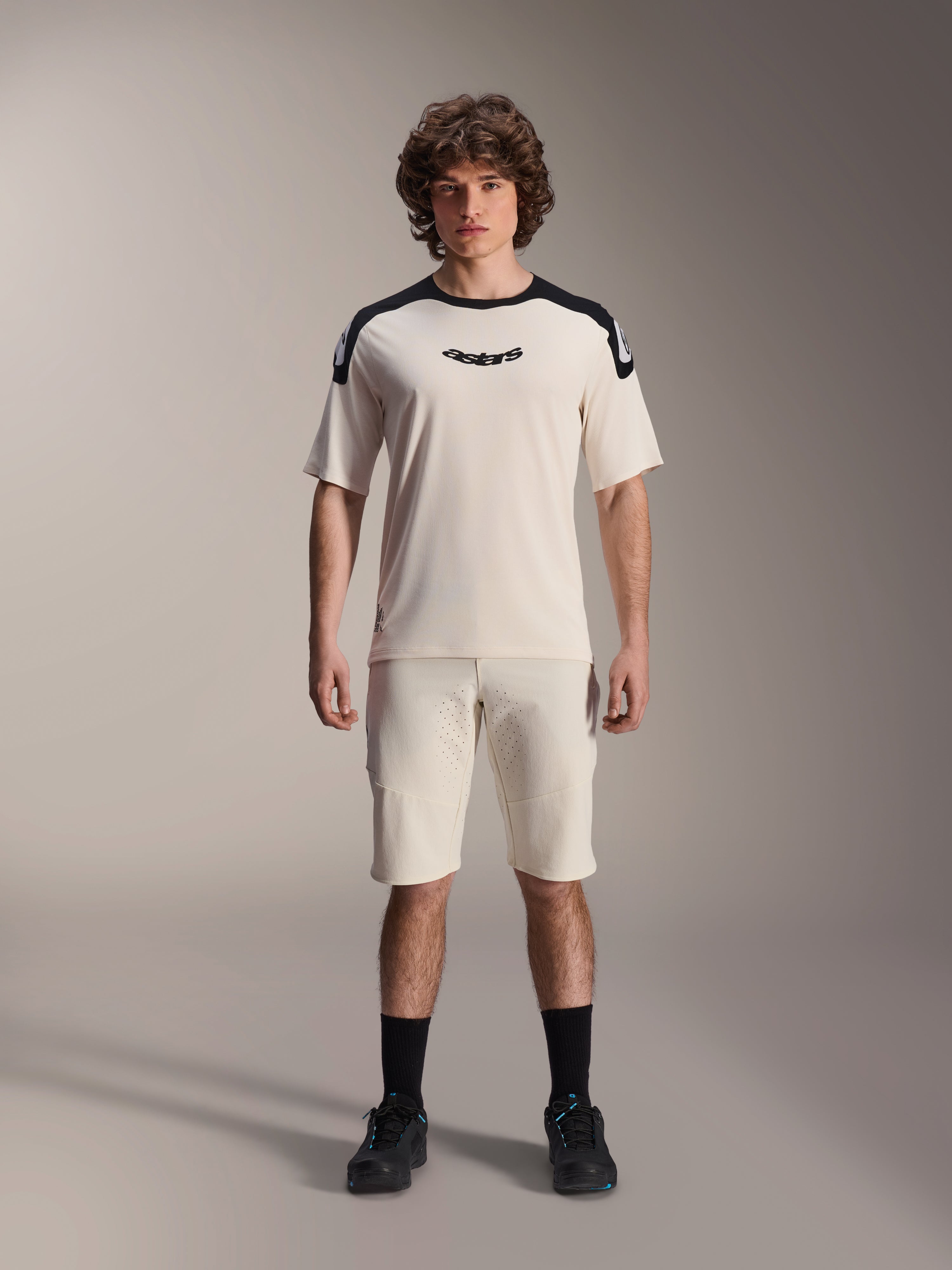 Alpinestars A-Aria Elite Shorts, pantaloncini da mountain bike, ecru, indossati con maglia a maniche corte coordinata con pannelli neri sulle spalle e logo, look sportivo casual con perforazioni frontali, abbigliamento tecnico da MTB