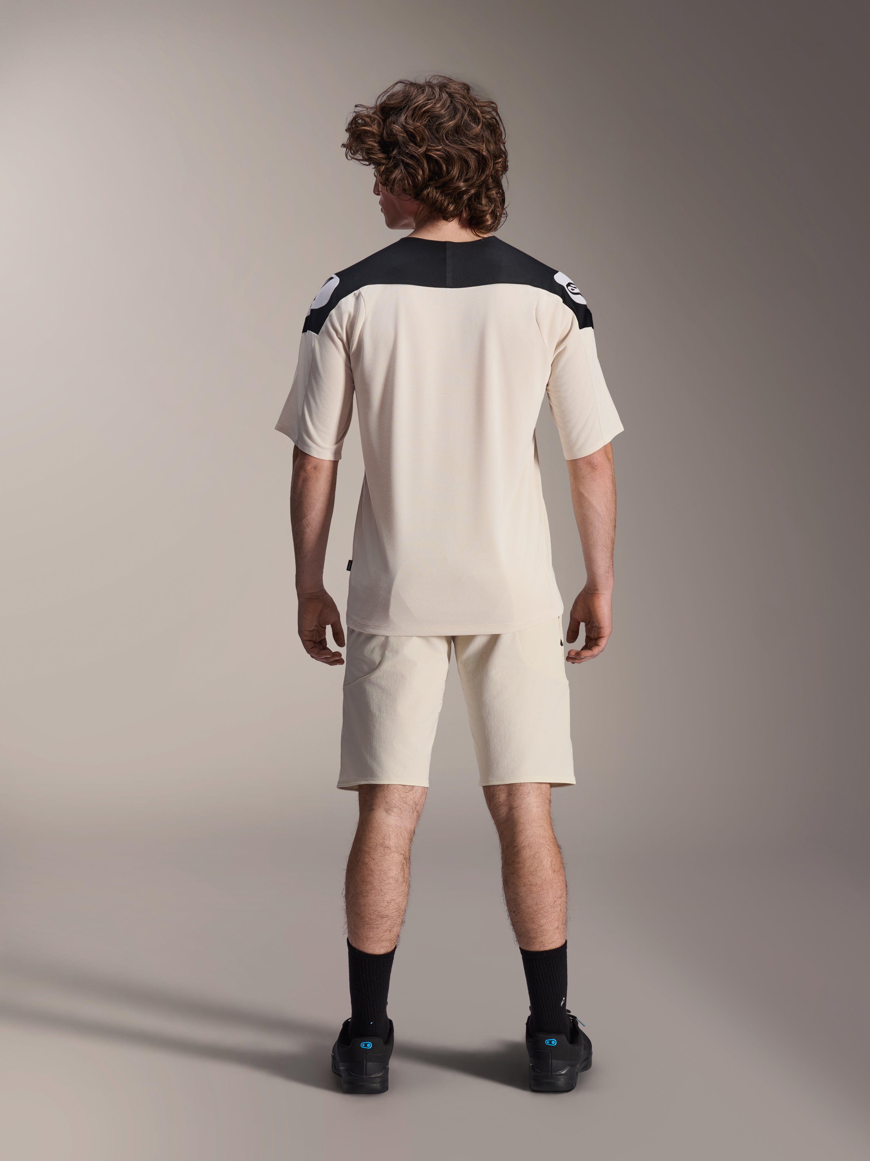 Maglia a maniche corte Alpinestars A-Aria Polartec Elite, maglia da ciclismo MTB, color ecru con pannelli sulle spalle neri, vista posteriore con pantaloncini coordinati e calze nere, vestibilità performante con design a coda allungata per la guida fuoristrada.