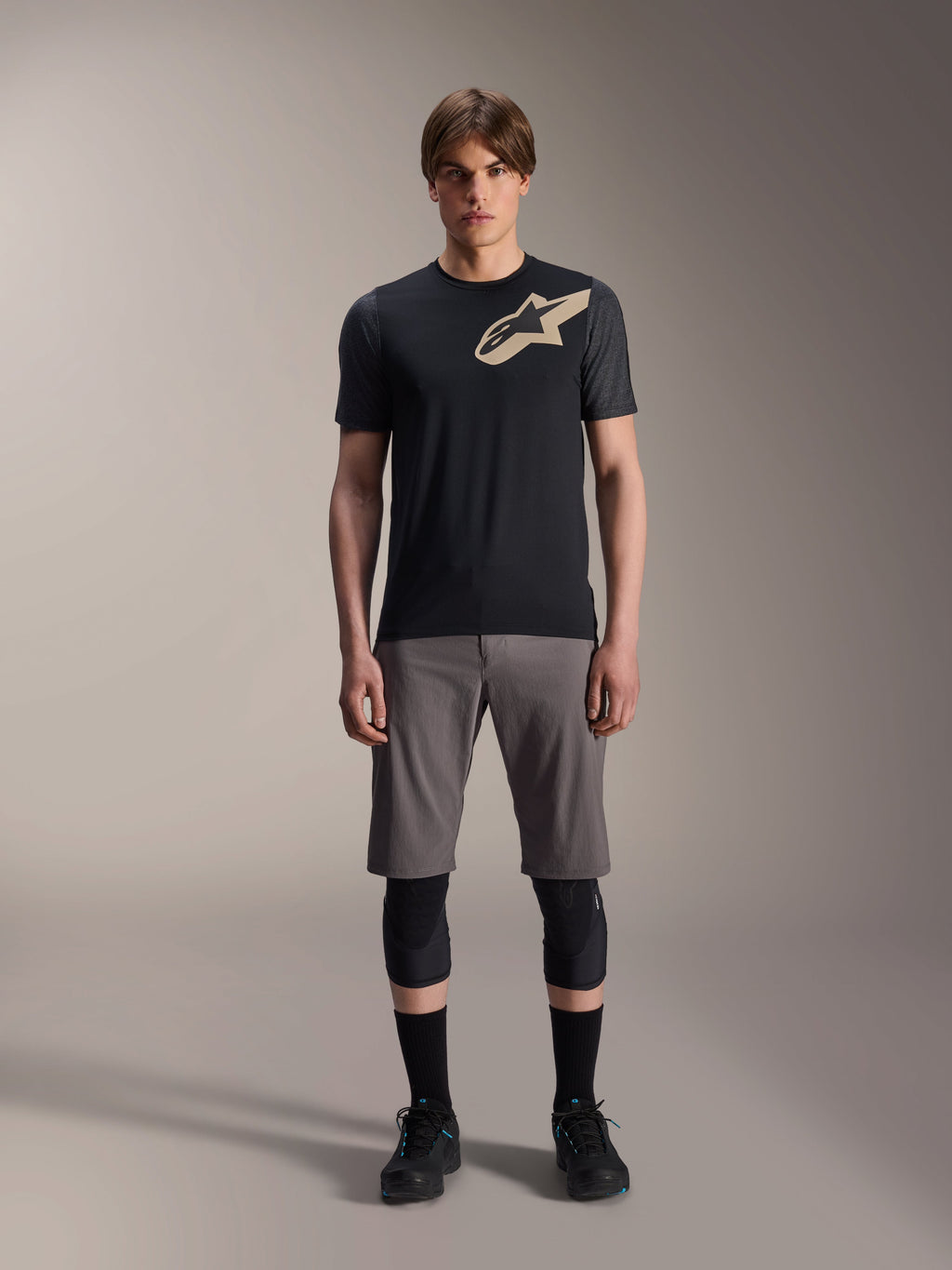 Alpinestars A-Dura Lipan Short Sleeve Jersey, maglia a maniche corte nera con maniche grigio melange e logo Astars color sabbia sul petto, indossata da un modello uomo con pantaloncini da mountain bike grigi e ginocchiere nere, abbigliamento tecnico da MTB