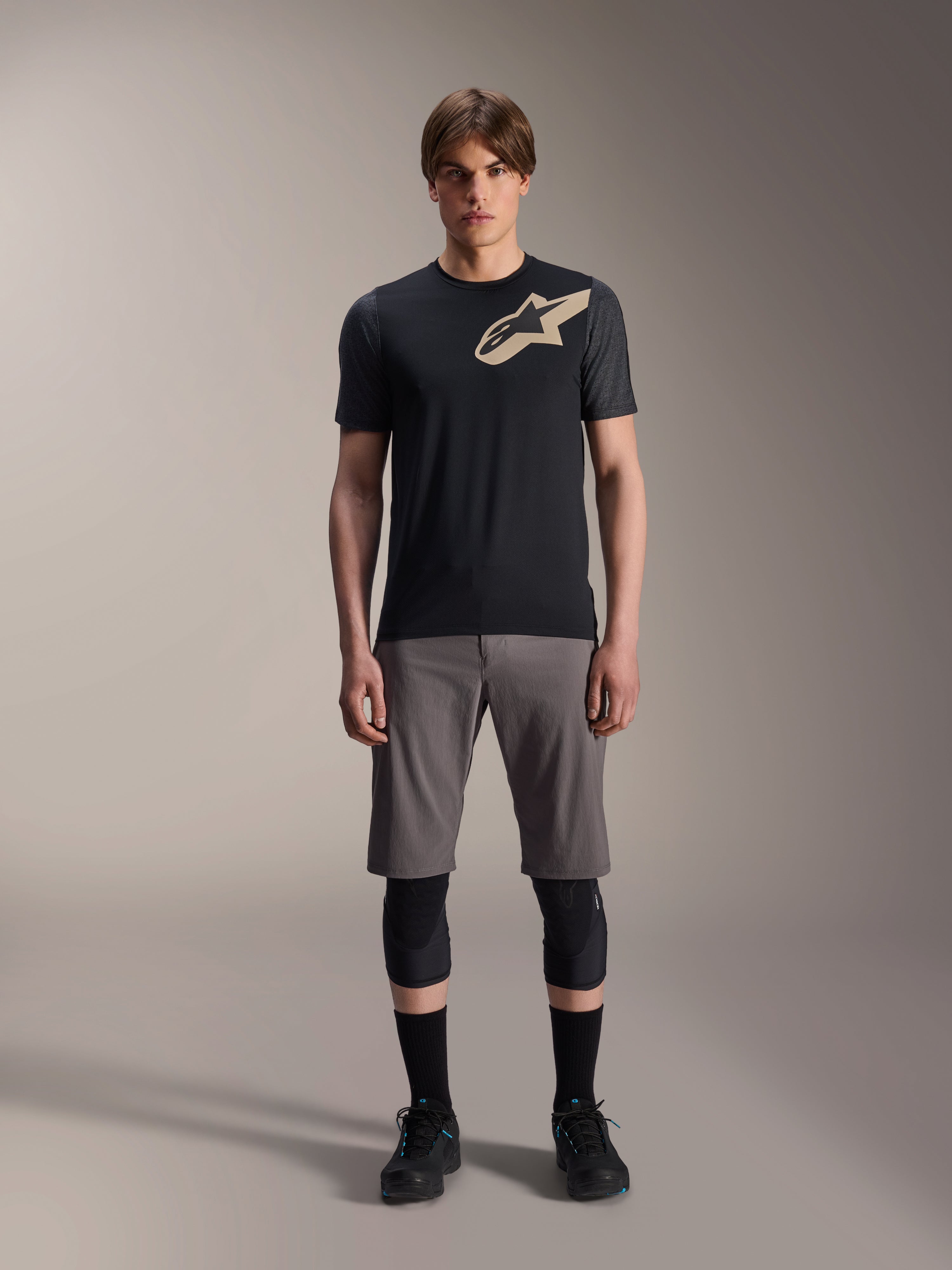 Alpinestars A-Dura Lipan Short Sleeve Jersey, maglia a maniche corte nera con maniche grigio melange e logo Astars color sabbia sul petto, indossata da un modello uomo con pantaloncini da mountain bike grigi e ginocchiere nere, abbigliamento tecnico da MTB