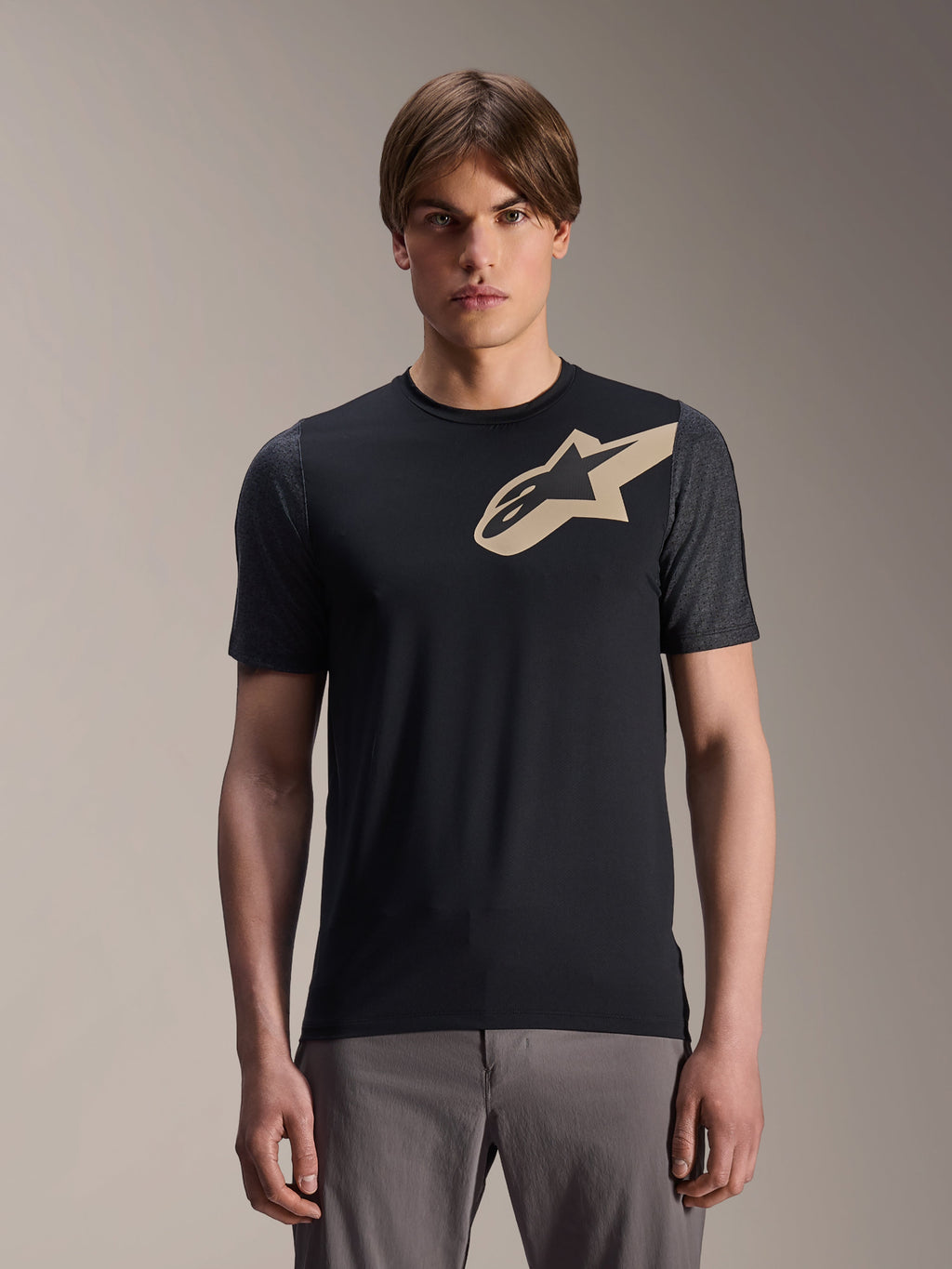 Modello che indossa la maglia Alpinestars A-Dura Lipan a maniche corte, maglia da MTB nera con logo a stella beige e maniche grigio scuro, abbinata a pantaloni da ciclismo grigi, abbigliamento tecnico casual