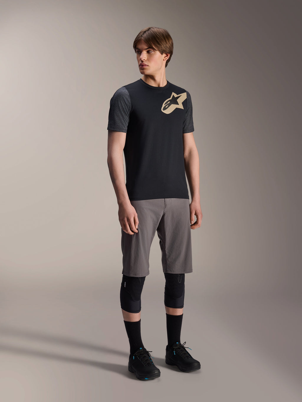 Modello uomo che indossa la maglia a maniche corte Alpinestars A-Dura Lipan nera, con maniche grigio scuro e logo Astars tan sul petto, abbinata a pantaloncini da mountain bike grigi, ginocchiere nere e scarpe da ciclismo nere, abbigliamento coordinato per mountain bike
