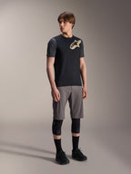 Modello uomo che indossa la maglia a maniche corte Alpinestars A-Dura Lipan nera, con maniche grigio scuro e logo Astars tan sul petto, abbinata a pantaloncini da mountain bike grigi, ginocchiere nere e scarpe da ciclismo nere, abbigliamento coordinato per mountain bike