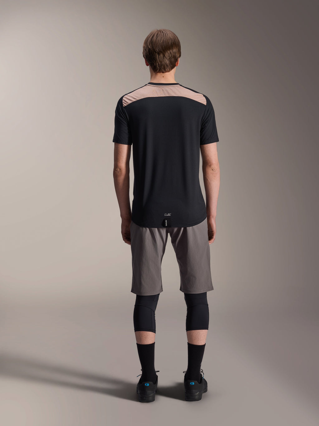 Maglia Alpinestars A-Dura Lipan - Maniche Corte, nera con pannello superiore posteriore color sabbia, vista posteriore con coda più lunga, indossata con pantaloncini MTB grigi e ginocchiere nere, abbigliamento tecnico da mountain bike