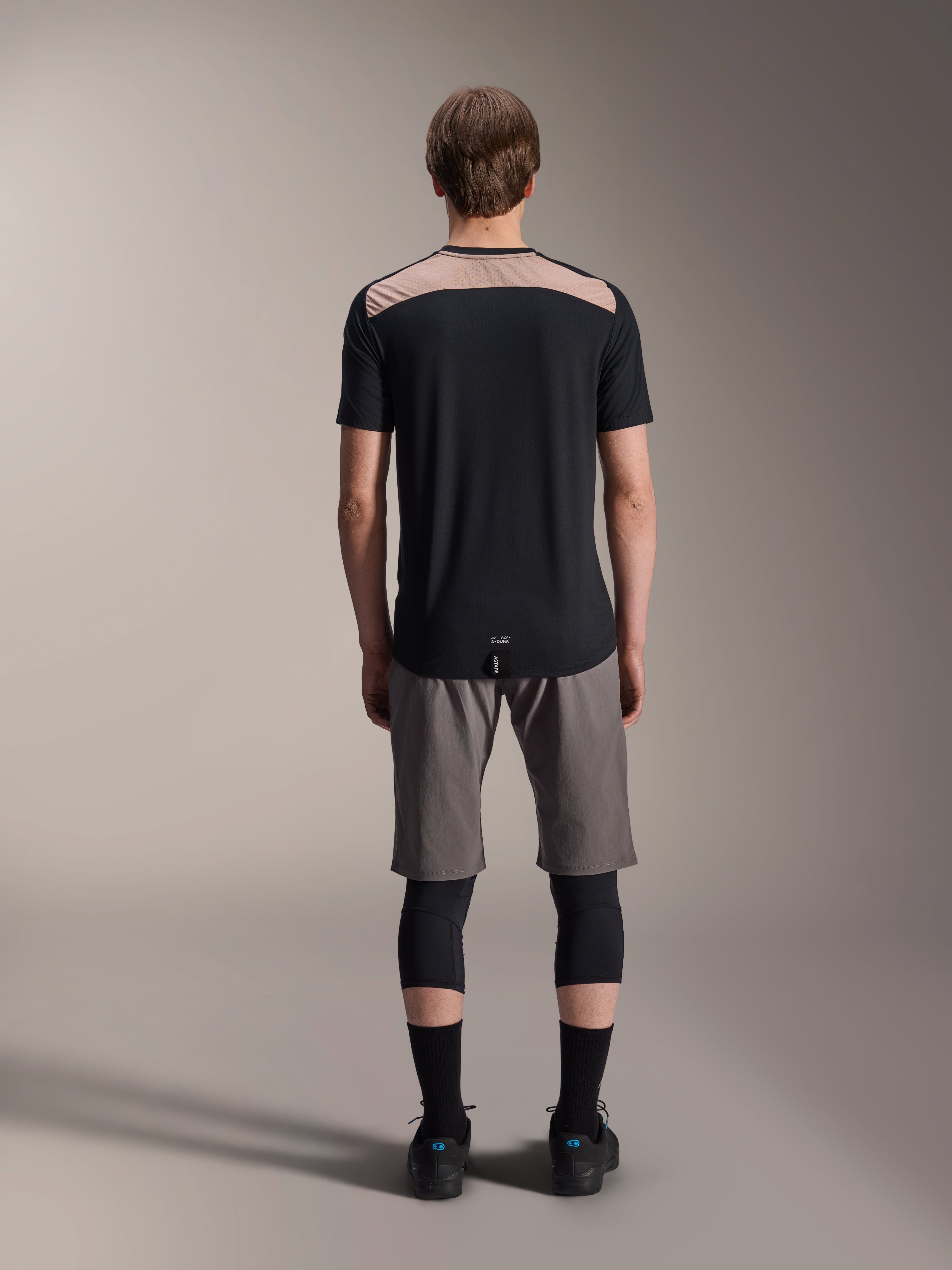 Maglia Alpinestars A-Dura Lipan - Maniche Corte, nera con pannello superiore posteriore color sabbia, vista posteriore con coda più lunga, indossata con pantaloncini MTB grigi e ginocchiere nere, abbigliamento tecnico da mountain bike