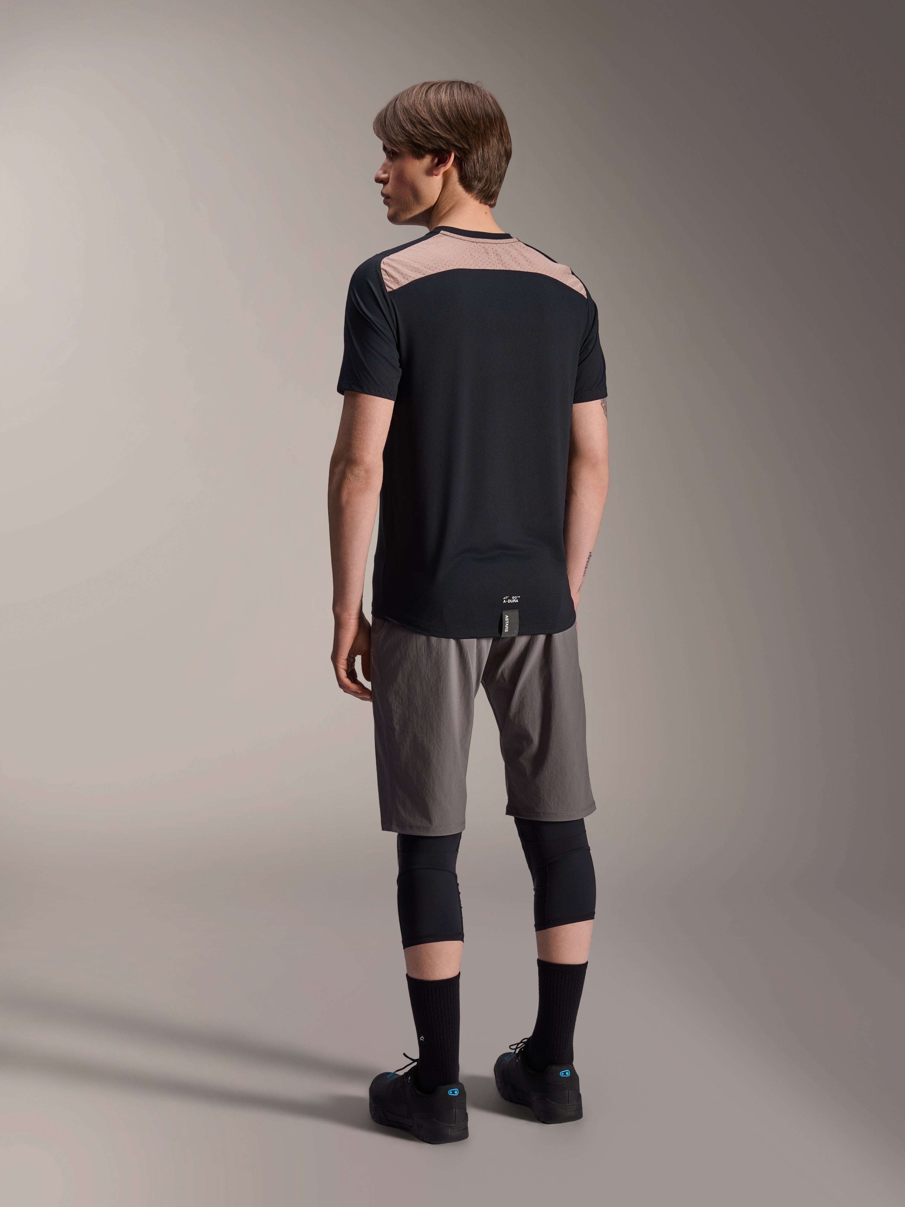 Maglia Alpinestars A-Dura Lipan a maniche corte in nero, vista posteriore su modello che mostra l'inserto in rete traforata marrone chiaro sulla parte superiore della schiena, abbinata a pantaloncini da mountain bike grigi e ginocchiere nere per un outfit completo da trail riding.