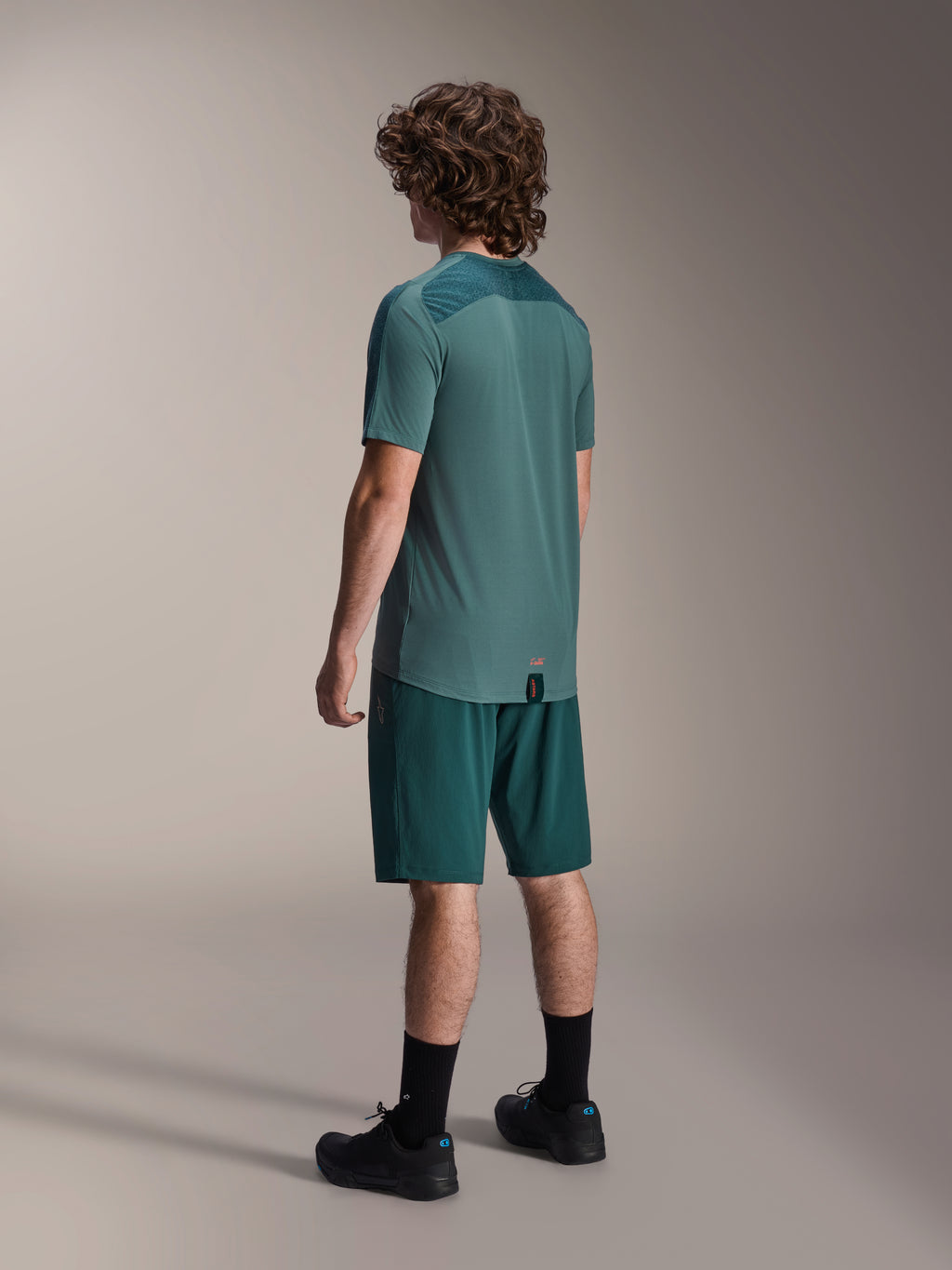 Modello che indossa i pantaloncini Alpinestars A-Dura in Dark Pine e la maglia a maniche corte Lipan verde coordinata, vista posteriore, abbinati a calze nere e scarpe da mountain bike nere, abbigliamento professionale da MTB.