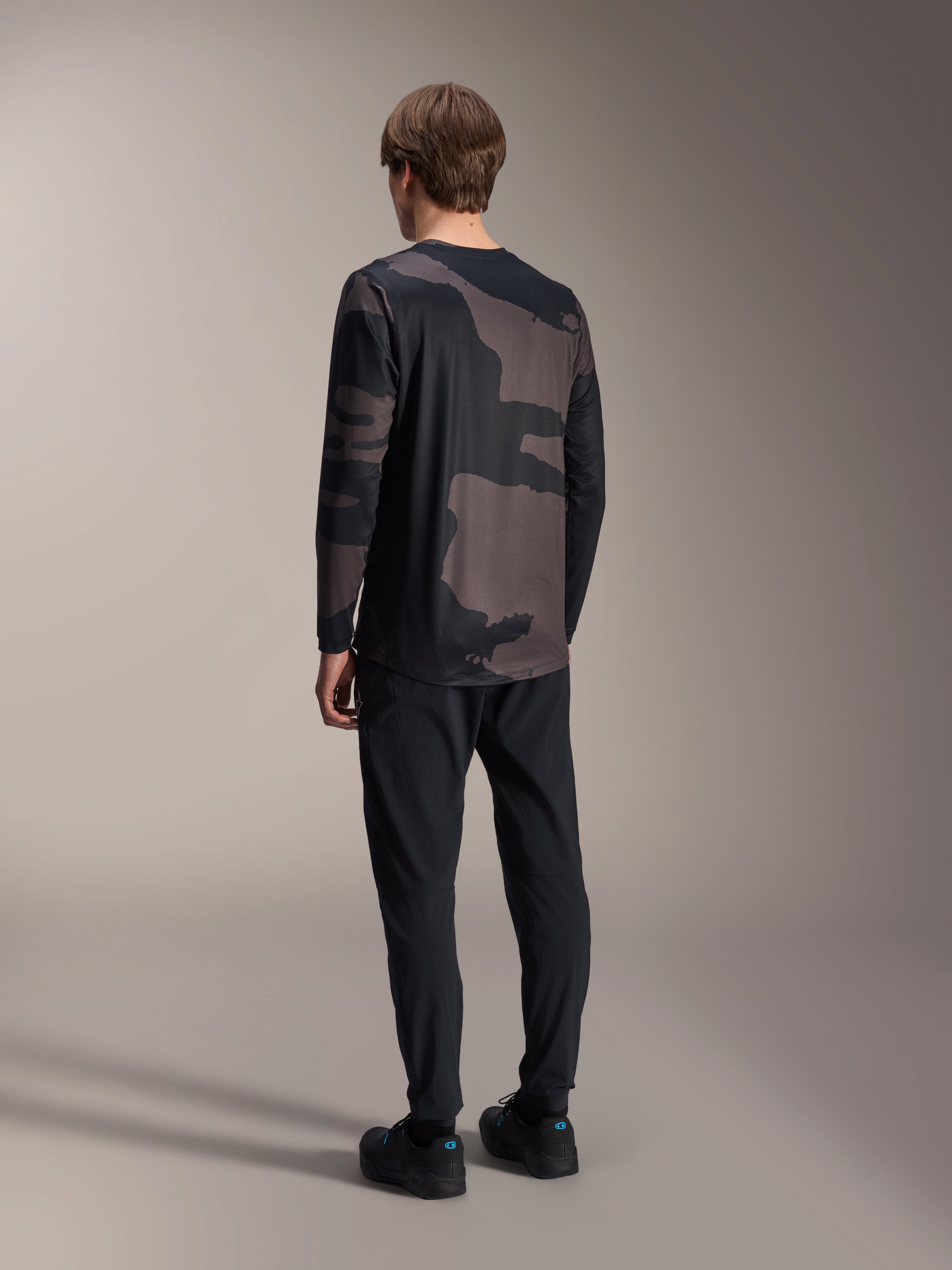 Alpinestars Maglia a maniche lunghe A-Dura Hollow Camo, Black Camo, vista posteriore del modello, maglia tecnica da mountain bike con orlo posteriore allungato, abbinata a pantaloni neri e scarpe da ciclismo, abbigliamento per prestazioni off-road dallo stile casual