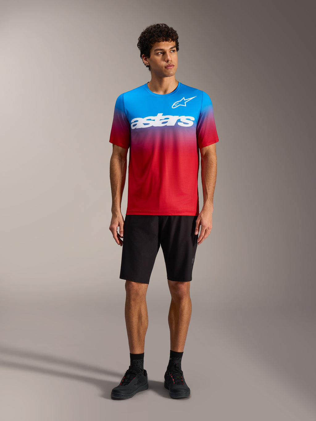 Maglia A-Dura ASTARS 2.0 con sfumatura rosso e blu, logo bianco, abbinata a pantaloncini e scarpe da MTB neri.