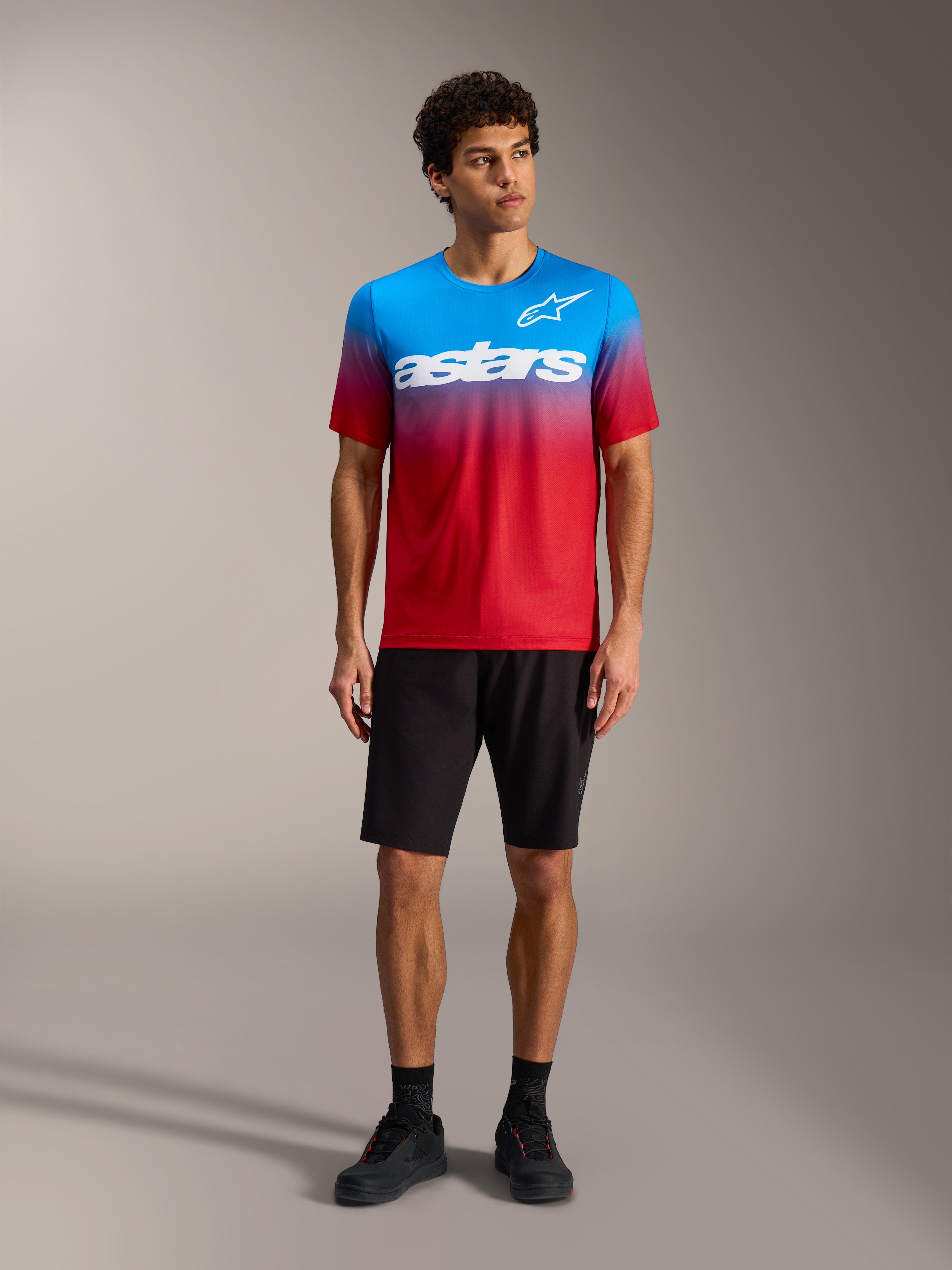 Maglia A-Dura ASTARS 2.0 con sfumatura rosso e blu, logo bianco, abbinata a pantaloncini e scarpe da MTB neri.