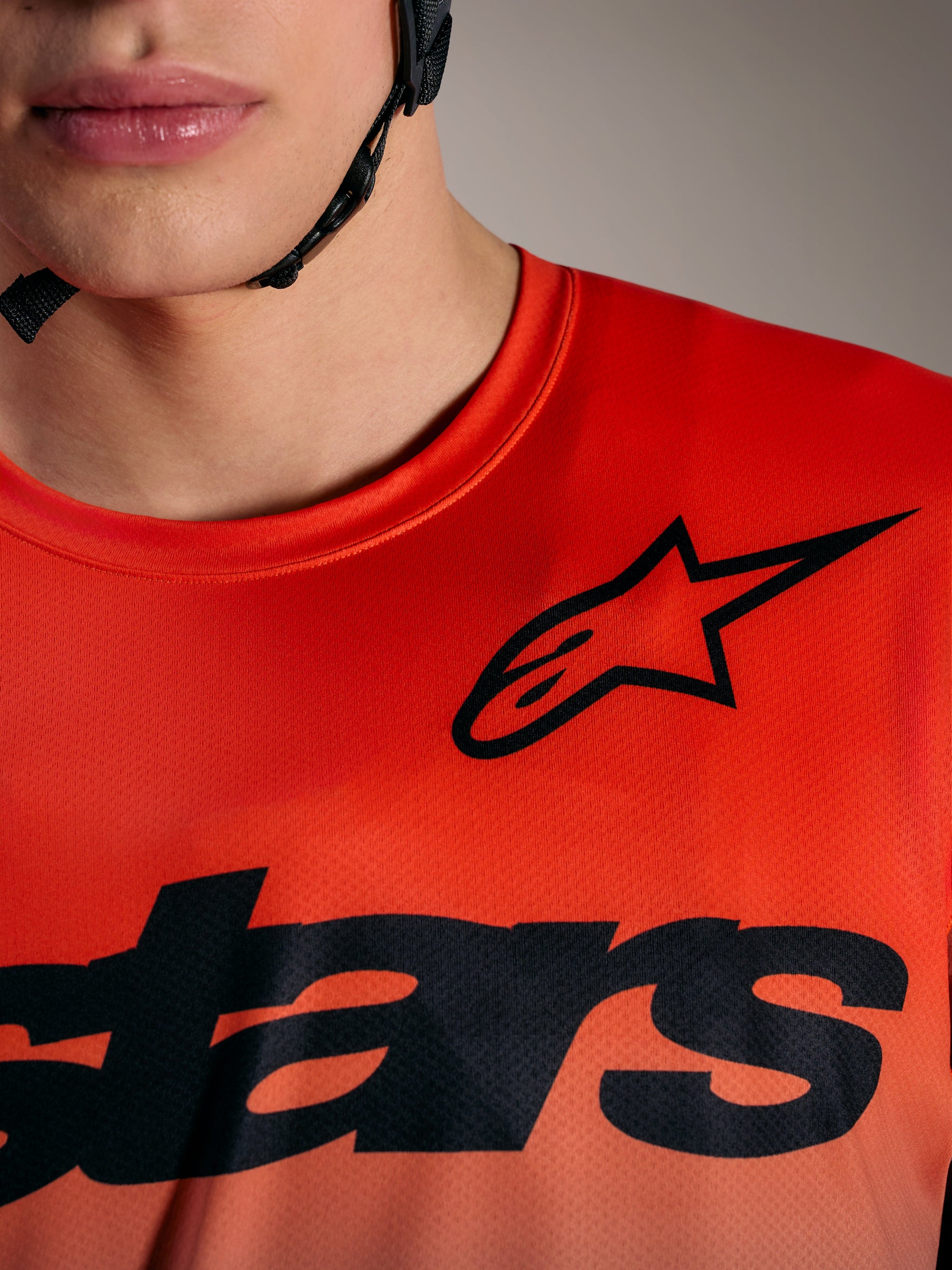 Maglia A-Dura ASTARS 2.0 arancione, primo piano dei loghi neri Alpinestars sul petto in tessuto leggero poly-spandex.