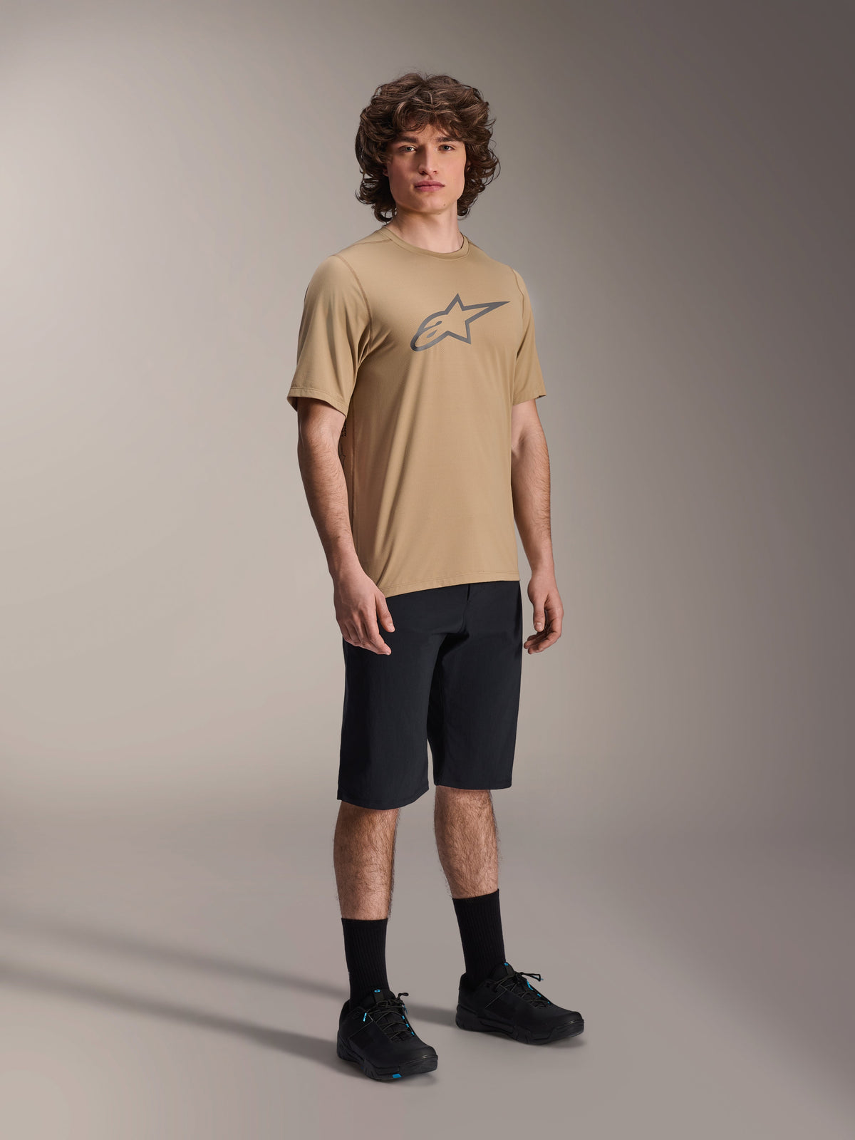 Maglia Alpinestars A-Dura Astar a maniche corte, marrone chiaro, con grande logo Astar grigio scuro sul petto, indossata da un modello con pantaloncini da MTB neri, calze e scarpe nere, abbigliamento tecnico casual da mountain bike in studio
