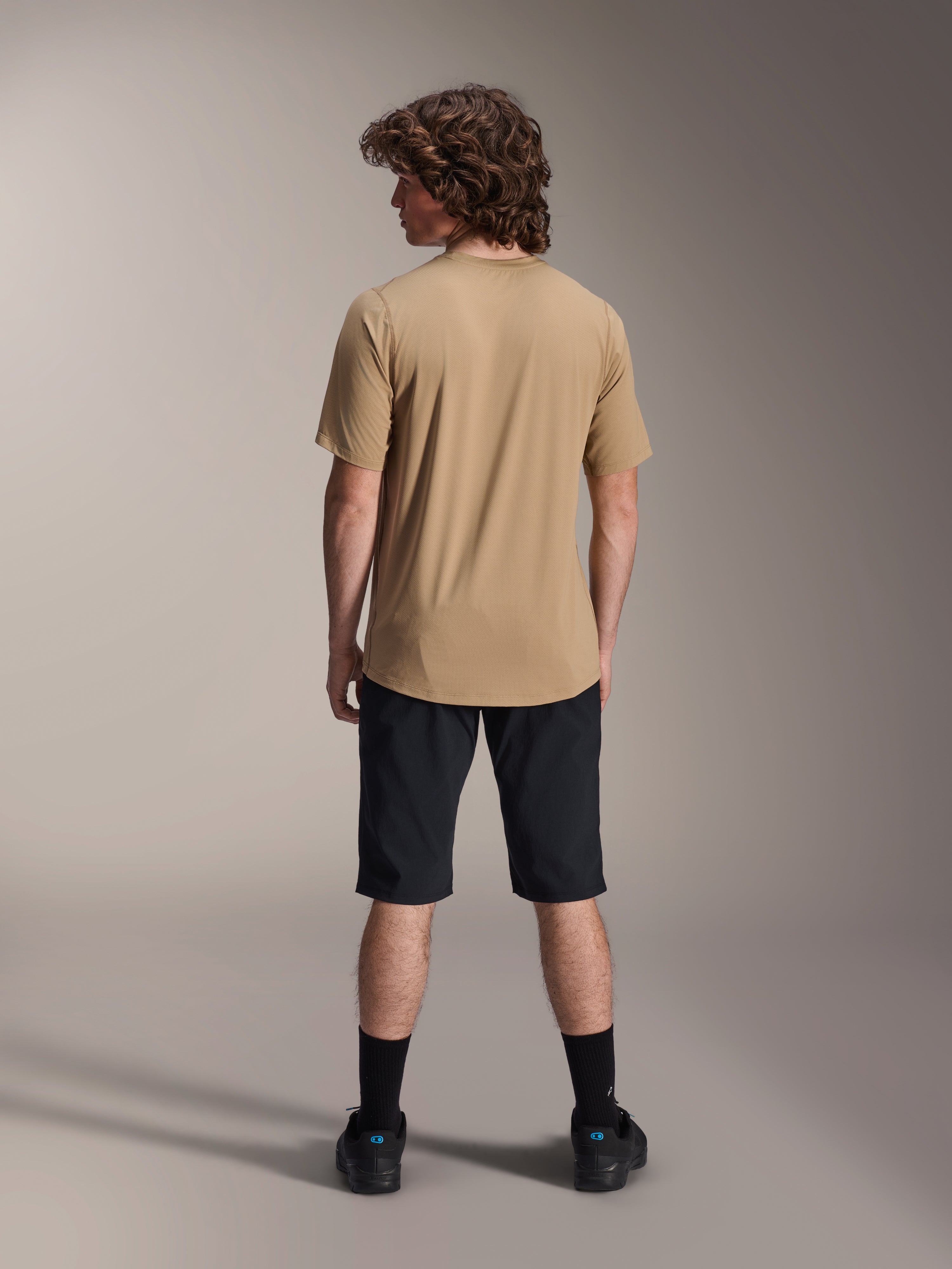 Modello maschile visto da dietro che indossa pantaloncini Alpinestars A-Dura Liner neri e una maglia a maniche corte color sabbia, abbinati a calze e scarpe nere, abbigliamento da mountain bike resistente per il ciclismo fuoristrada.