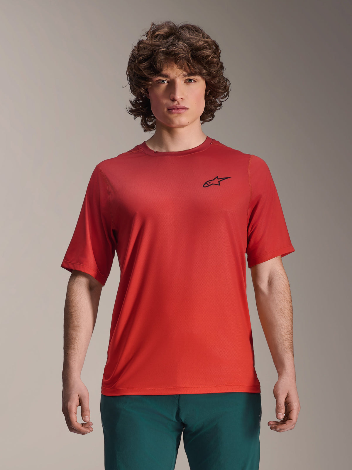 Alpinestars 2025 A-Dura Rise Jersey, maglia MTB a maniche corte, Spicy Orange, indossata da un uomo, con logo Astars nero sul petto, abbinata a pantaloni verde scuro, abbigliamento tecnico casual per mountain bike
