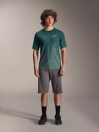 Modello che indossa la maglia a maniche corte Alpinestars A-Dura Rise in Silver Pine, maglia da ciclismo verde con piccolo logo bianco sul petto, abbinata a pantaloncini da mountain bike grigi e calzature nere, abbigliamento casual da trail MTB
