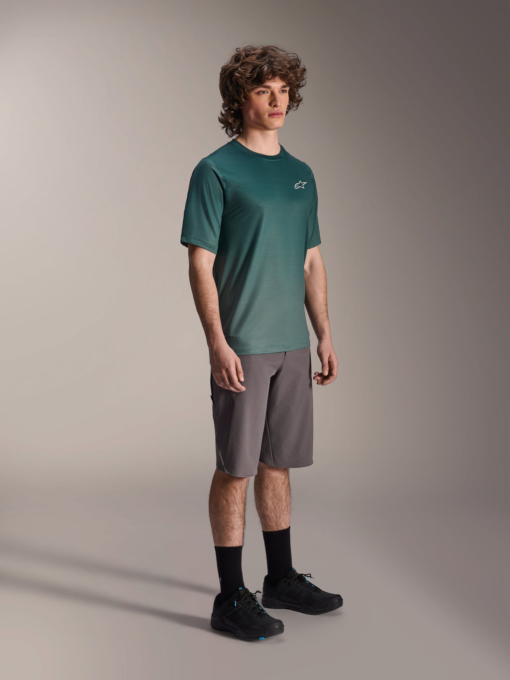 Alpinestars Maglia A-Dura Rise - Manica Corta in Silver Pine, maglia da mountain bike verde indossata da un modello con pantaloncini grigi e sneakers nere, abbigliamento casual per trail riding MTB con un piccolo logo bianco sul petto