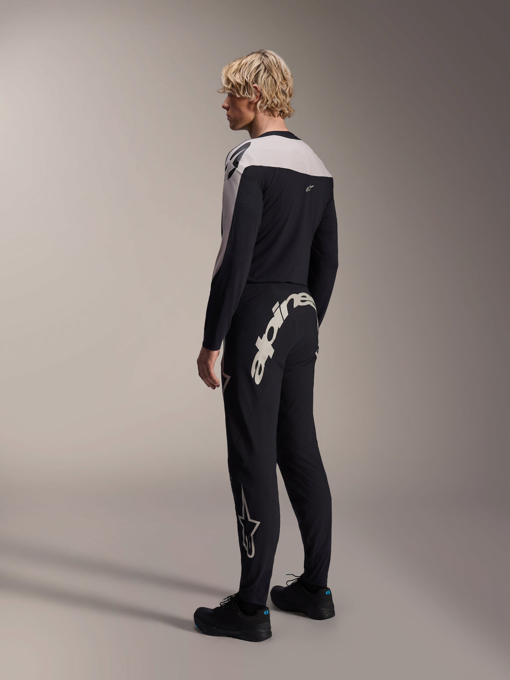 Alpinestars A-Supra Melt Pants, pantaloni da MTB, nero, caratterizzati da una grande scritta Alpinestars bianca sul carré posteriore e logo a stella sulla gamba, indossati da un modello con maglia a maniche lunghe coordinata e scarpe nere, vista posteriore laterale
