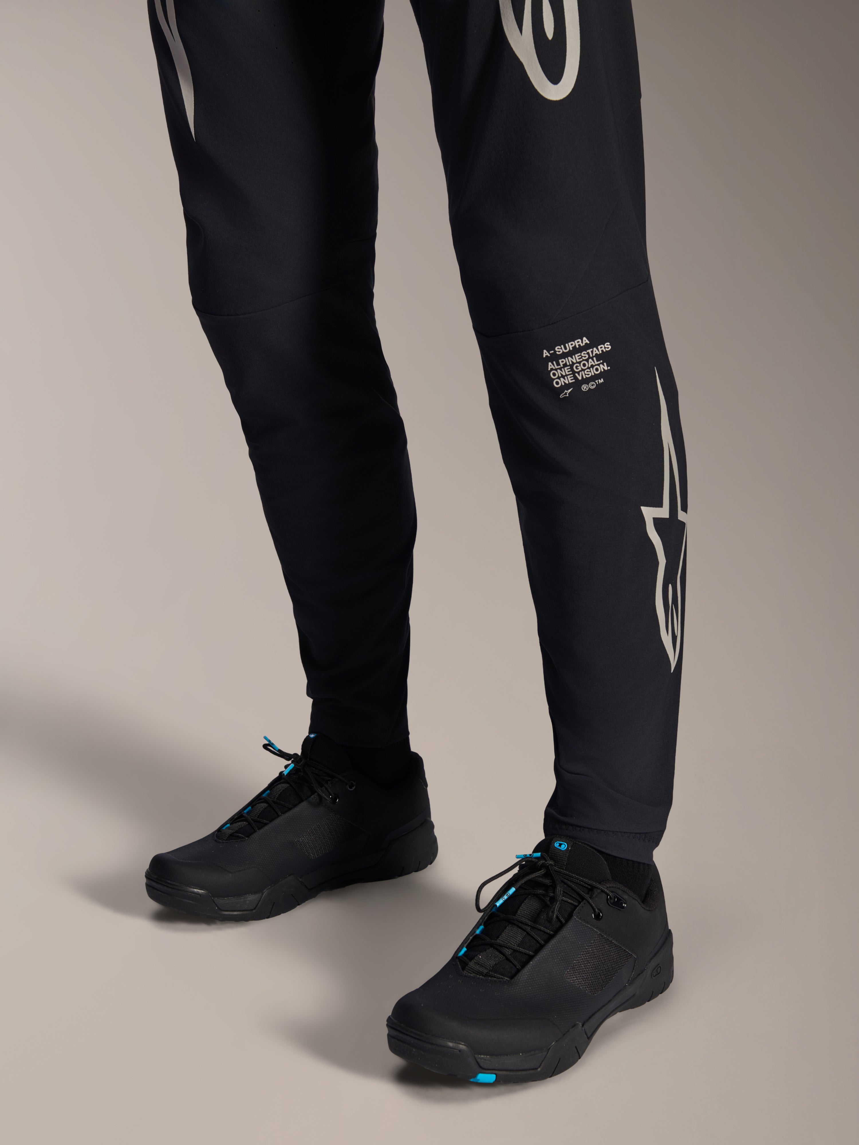 Pantaloni Alpinestars A-Supra Melt, pantaloni da mountain bike neri, con scritta e logo Alpinestars bianchi sulla gamba, indossati con scarpe da ciclismo nere con accenti blu, abbigliamento tecnico per fuoristrada