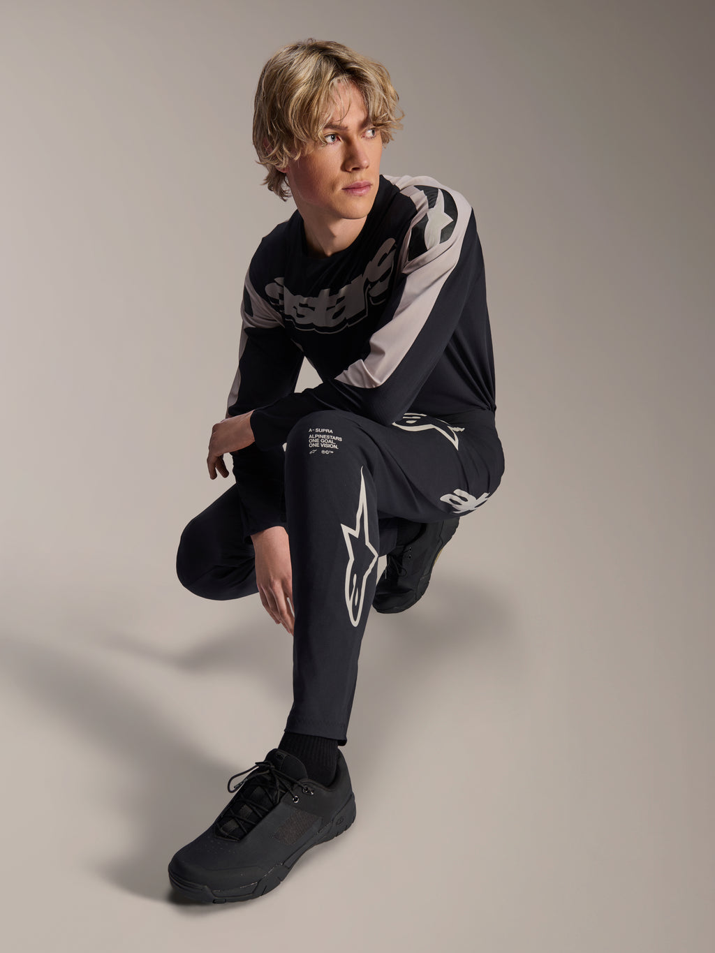 Pantaloni e maglia a maniche lunghe Alpinestars A-Supra Melt, nero e grigio, caratterizzati da audace branding bianco e loghi a stella, indossati da un modello maschile in posa accovacciata, completo coordinato da gara MTB.