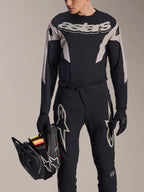 Maglia Alpinestars A-Supra Melt a maniche lunghe, nero con pannelli grafici grigi e vistosa scritta 'astars' sul petto, indossata con pantaloni e guanti da mountain bike neri mentre tiene un casco integrale nero, kit da gara MTB performante