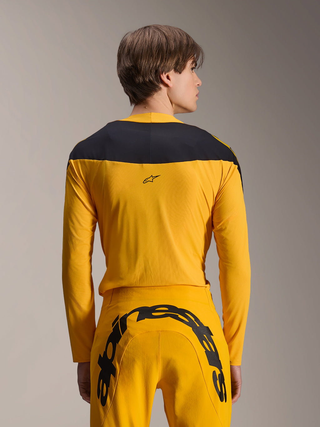 Alpinestars Limited Edition A-Supra Melt Gold Gear-Set, set abbigliamento MTB giallo, vista posteriore del modello che indossa una maglia a maniche lunghe con pannello nero sulle spalle e pantaloni gialli coordinati con un grande logo nero sul retro, abbigliamento da ciclismo ad alte prestazioni.