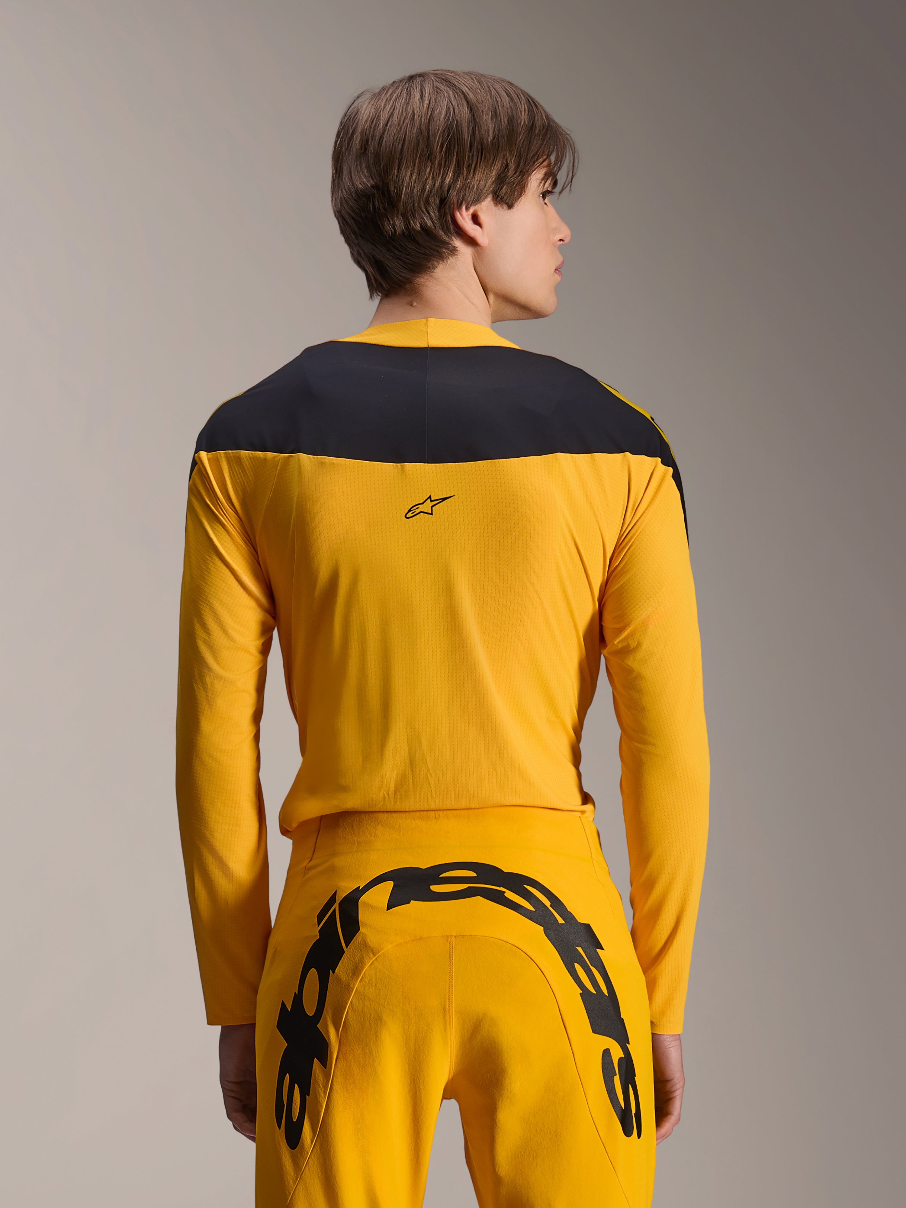 Alpinestars Limited Edition A-Supra Melt Gold Gear-Set, set abbigliamento MTB giallo, vista posteriore del modello che indossa una maglia a maniche lunghe con pannello nero sulle spalle e pantaloni gialli coordinati con un grande logo nero sul retro, abbigliamento da ciclismo ad alte prestazioni.
