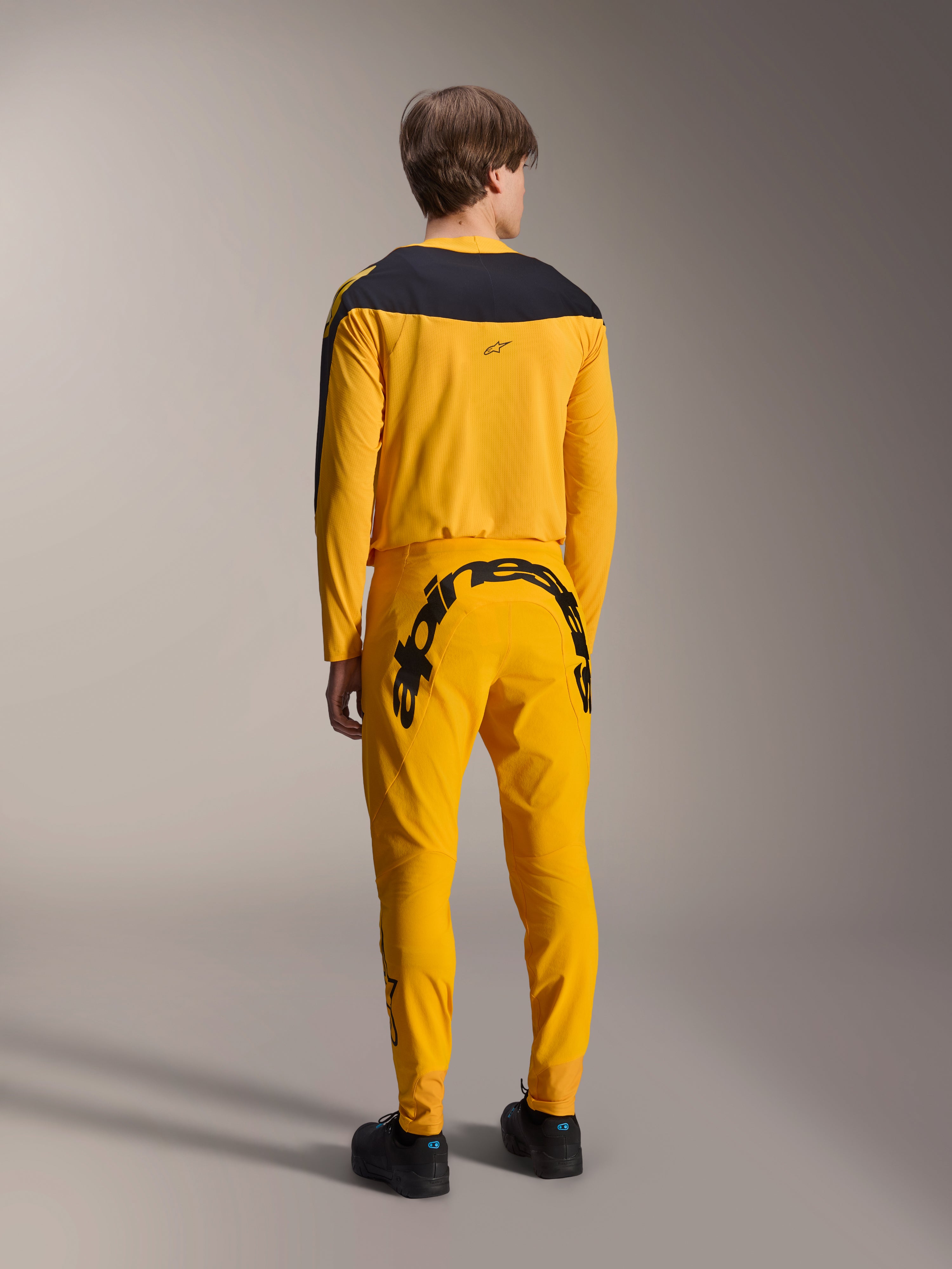 Alpinestars Maglia A-Supra Melt Gold Edizione Limitata, maglia MTB a maniche lunghe, giallo scuro con pannello nero sulle spalle, indossata con pantaloni gialli coordinati con vistoso marchio nero sul retro, vista posteriore di kit da ciclismo ad alte prestazioni