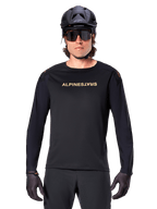 Maglia a maniche lunghe Alpinestars A-Aria Polartec Switch, nero, logo Alpinestars dorato sul petto, indossata con un casco da MTB nero, occhiali da sole scuri e guanti neri, abbigliamento professionale da trail per mountain bike