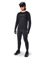 Alpinestars A-Aria Polartec Switch Jersey a maniche lunghe, nero, logo bronzo sul petto, indossata da un mountain biker con casco nero e occhiali da sole, abbigliamento tecnico da ciclismo per trail riding