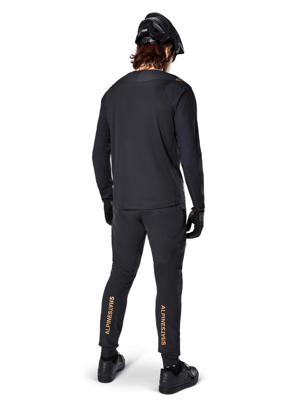 Vista posteriore di un rider di mountain bike che indossa la maglia a maniche lunghe Alpinestars A-Aria Polartec Switch in nero, abbinata a pantaloni da MTB neri con branding Alpinestars color sabbia sui polpacci, casco e guanti neri, equipaggiamento professionale da ciclismo per percorsi fuoristrada.