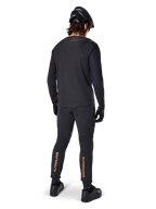 Vista posteriore di un rider di mountain bike che indossa la maglia a maniche lunghe Alpinestars A-Aria Polartec Switch in nero, abbinata a pantaloni da MTB neri con branding Alpinestars color sabbia sui polpacci, casco e guanti neri, equipaggiamento professionale da ciclismo per percorsi fuoristrada.