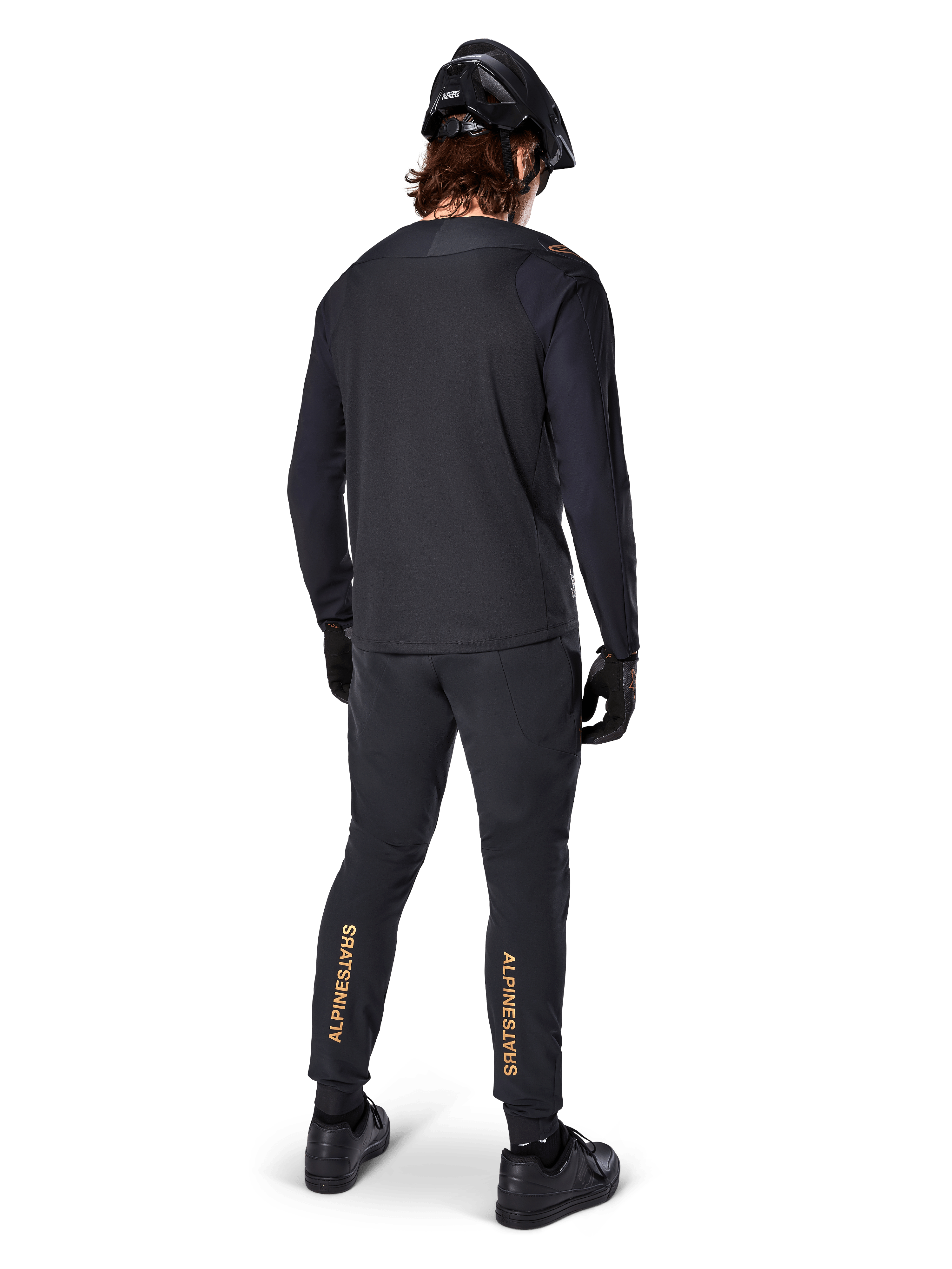 Vista posteriore di un rider di mountain bike che indossa la maglia a maniche lunghe Alpinestars A-Aria Polartec Switch in nero, abbinata a pantaloni da MTB neri con branding Alpinestars color sabbia sui polpacci, casco e guanti neri, equipaggiamento professionale da ciclismo per percorsi fuoristrada.