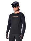 Alpinestars Maglia A-Aria Polartec Switch a maniche lunghe, nero con logo oro, abbigliamento per mountain bike MTB, indossata con casco nero e occhiali da sole, con ventilazione tagliata al laser sulle maniche e tessuto Polartec traspirante per prestazioni off-road