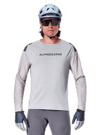 Alpinestars A-Aria Polartec Switch Long Sleeve Jersey, maglia da mountain bike grigio chiaro con perforazioni laser sulle maniche e logo sul petto, indossata con casco celeste, occhiali da sole e guanti grigi