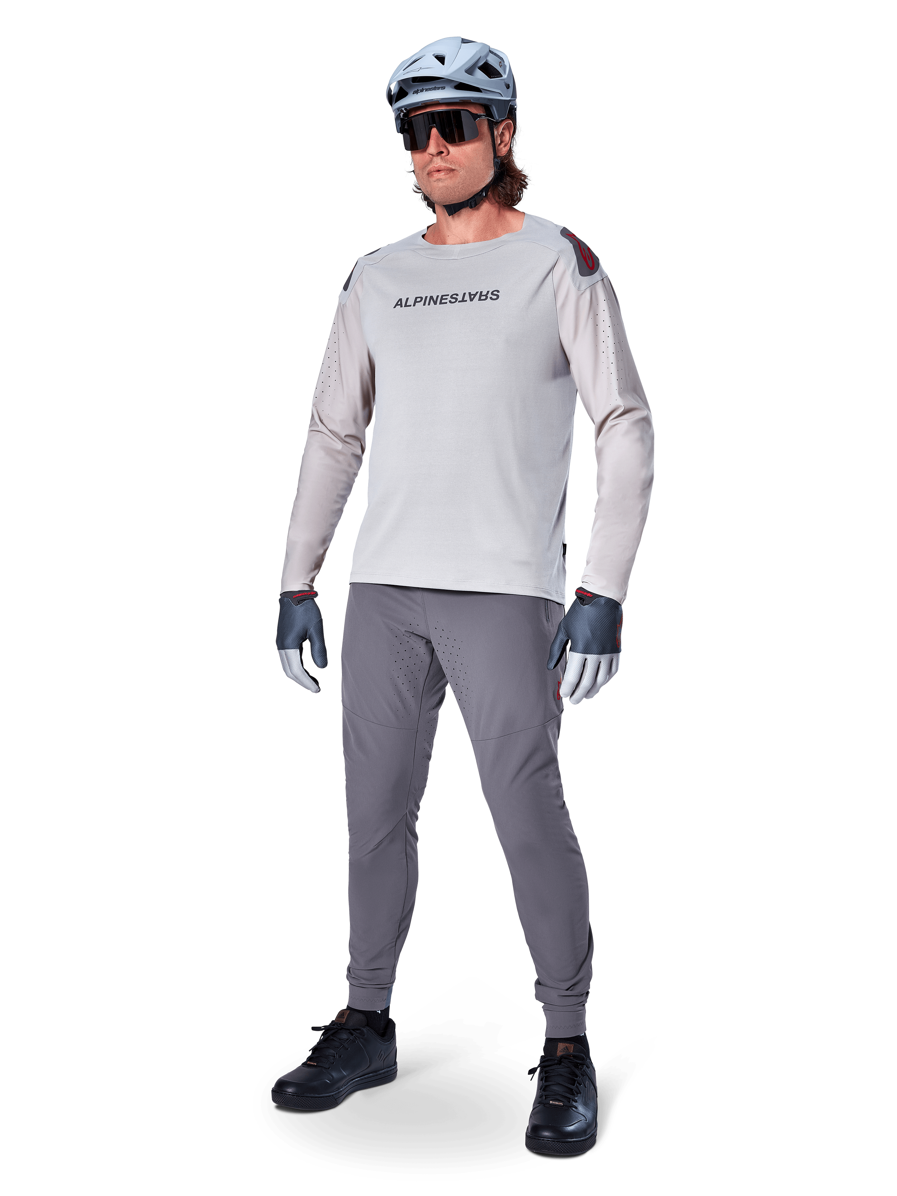 Alpinestars A-Aria Polartec Switch Jersey, maglia da MTB a maniche lunghe, grigio chiaro, indossata con pantaloni da mountain bike grigio scuro, casco e guanti, con maniche traforate al laser e marchio Alpinestars sul petto.