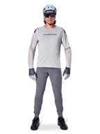 Maglia Alpinestars A-Aria Polartec Switch, a maniche lunghe, grigio chiaro, indossata da un ciclista di mountain bike con pantaloni MTB grigio scuro, casco azzurro chiaro e occhiali da sole, con marchio sul petto e ventilazione traforata al laser sulle maniche, outfit completo da riding off-road.