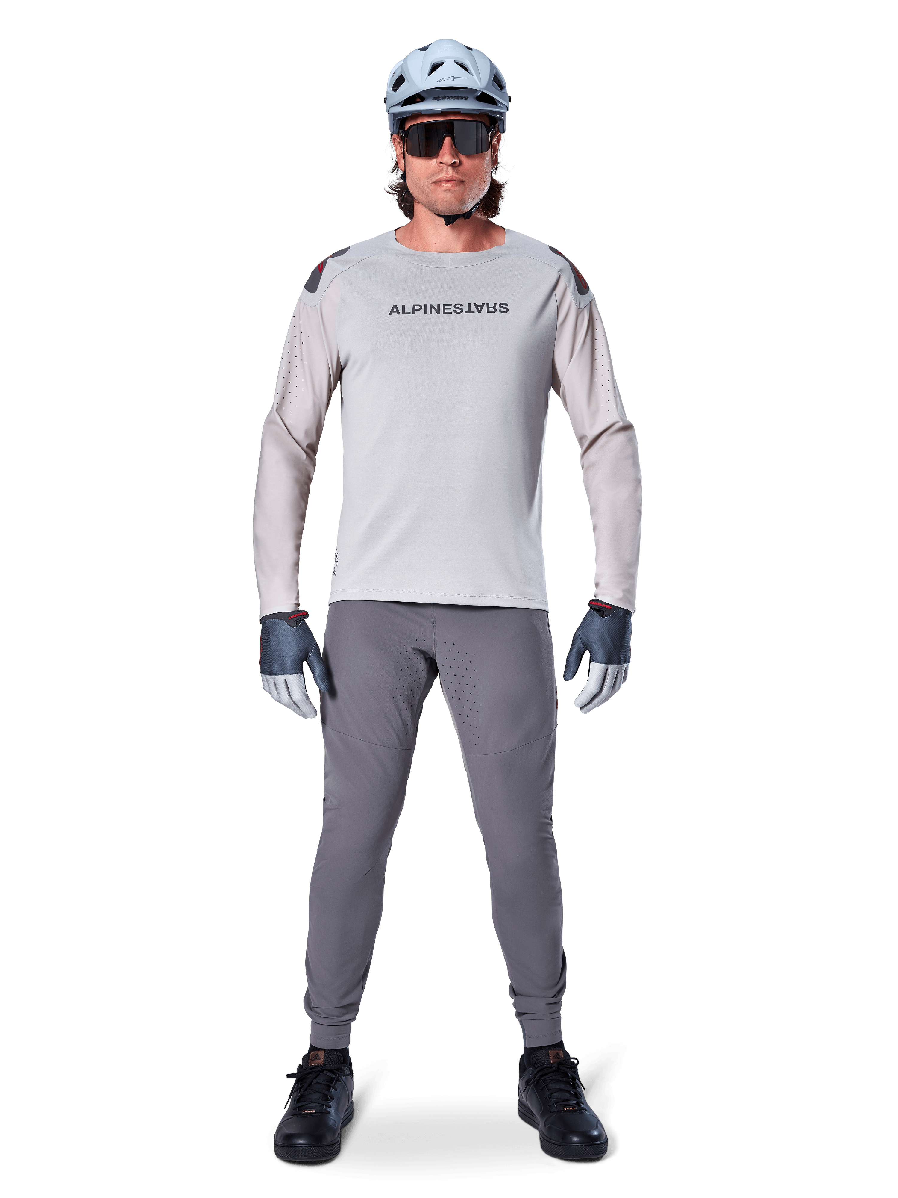 Maglia Alpinestars A-Aria Polartec Switch, a maniche lunghe, grigio chiaro, indossata da un ciclista di mountain bike con pantaloni MTB grigio scuro, casco azzurro chiaro e occhiali da sole, con marchio sul petto e ventilazione traforata al laser sulle maniche, outfit completo da riding off-road.