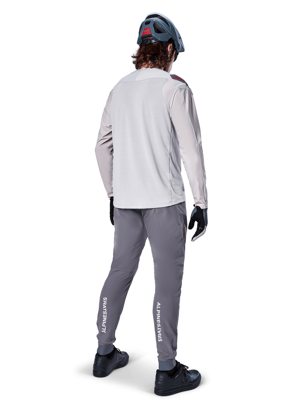 Vista posteriore di un ciclista che indossa la maglia a maniche lunghe Alpinestars A-Aria Polartec Switch in grigio chiaro, abbinata a pantaloni da mountain bike grigio scuro con marchio Alpinestars bianco sui polpacci, guanti neri e un casco grigio