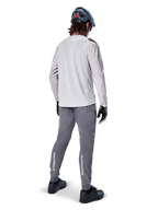 Vista posteriore di un ciclista che indossa la maglia a maniche lunghe Alpinestars A-Aria Polartec Switch in grigio chiaro, abbinata a pantaloni da mountain bike grigio scuro con marchio Alpinestars bianco sui polpacci, guanti neri e un casco grigio